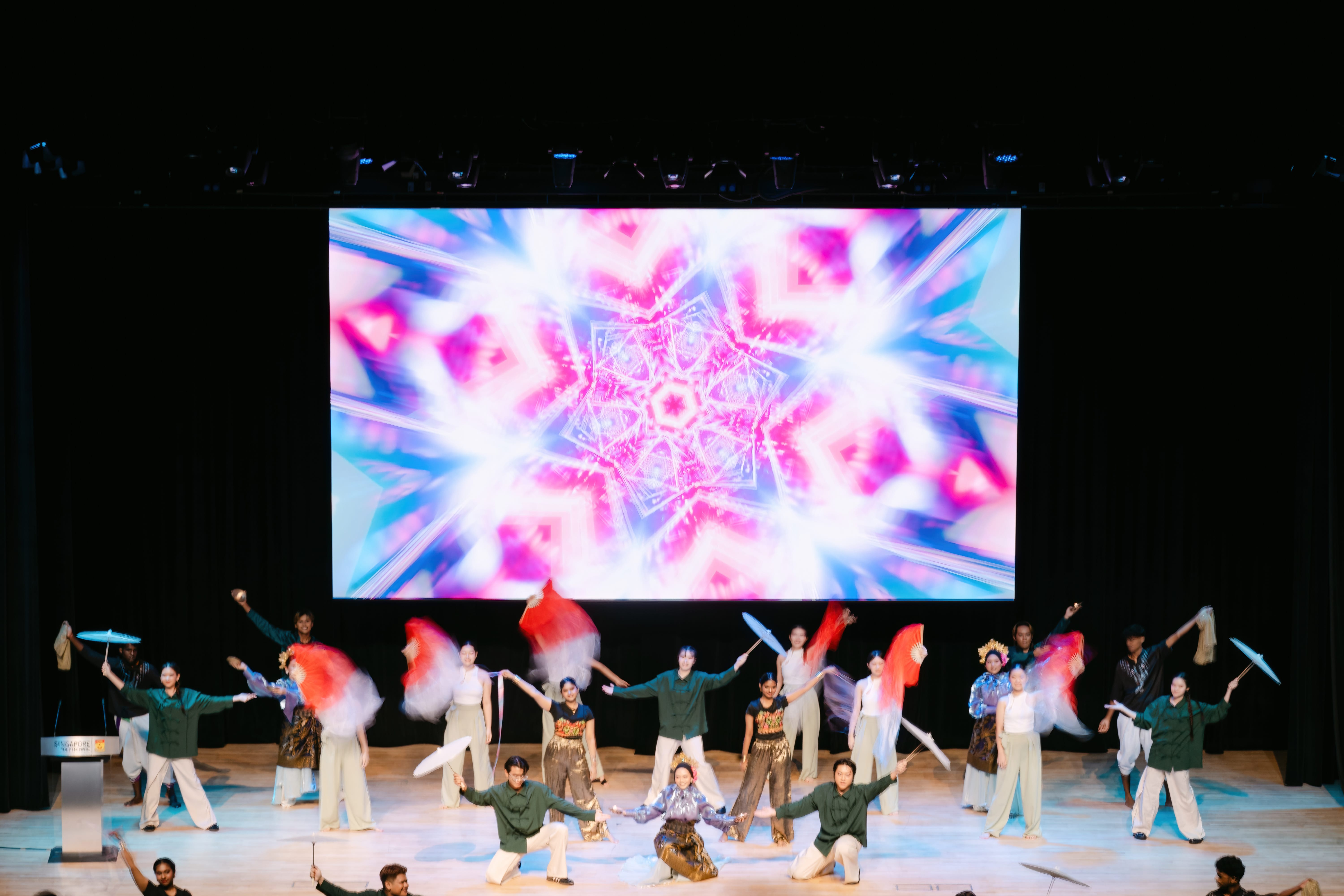 Performers in colorful costumes dance on a decorated stage with parasols and ribbons, backed by a vivid LED screen.