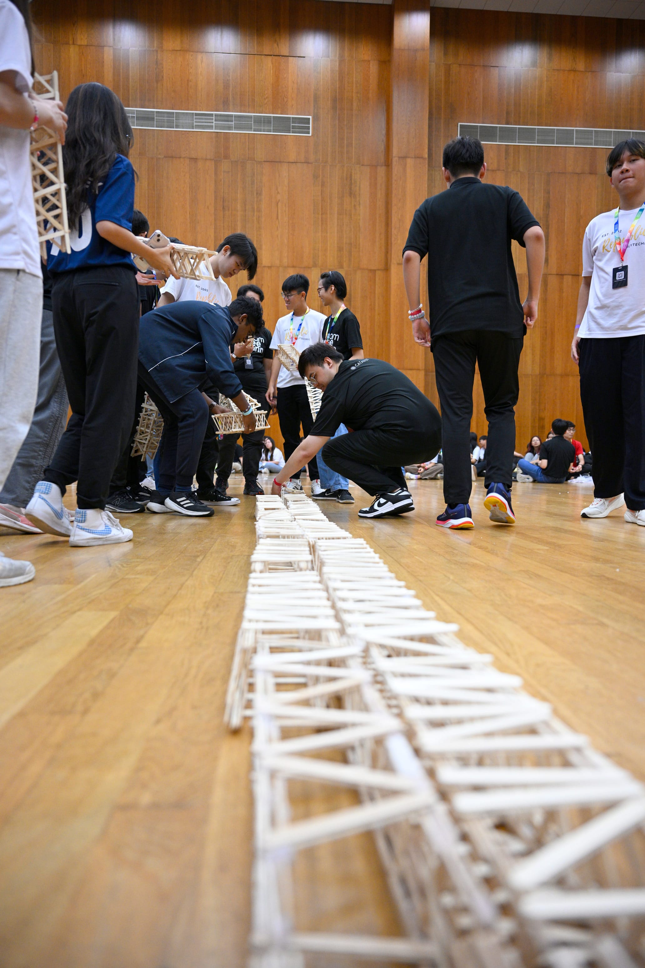 Students assemble a long line of small wooden stick bridge structures on a polished wooden hall floor.