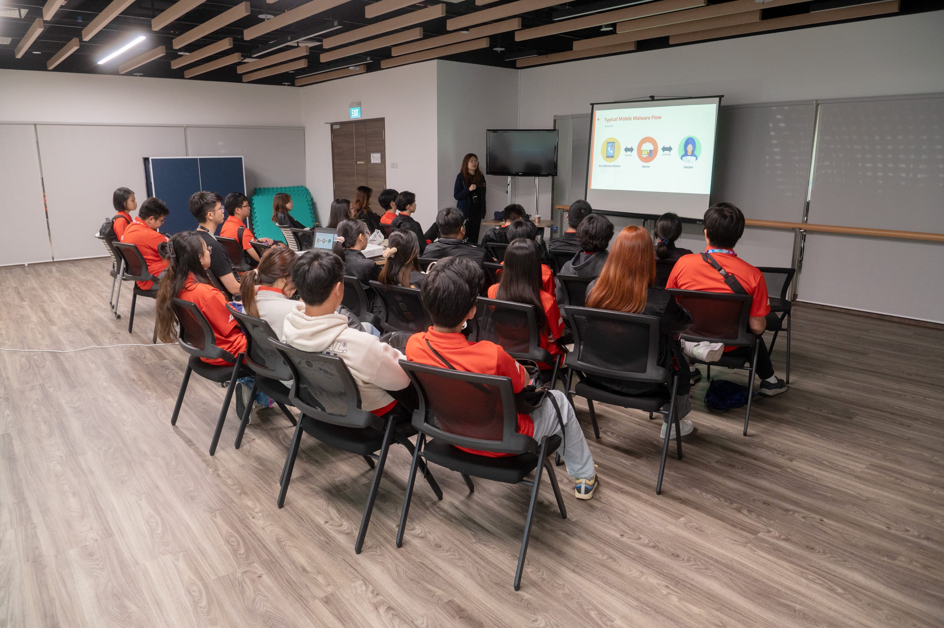 A presenter stands before a projected slide on mobile malware while students in red shirts sit in chairs listening.