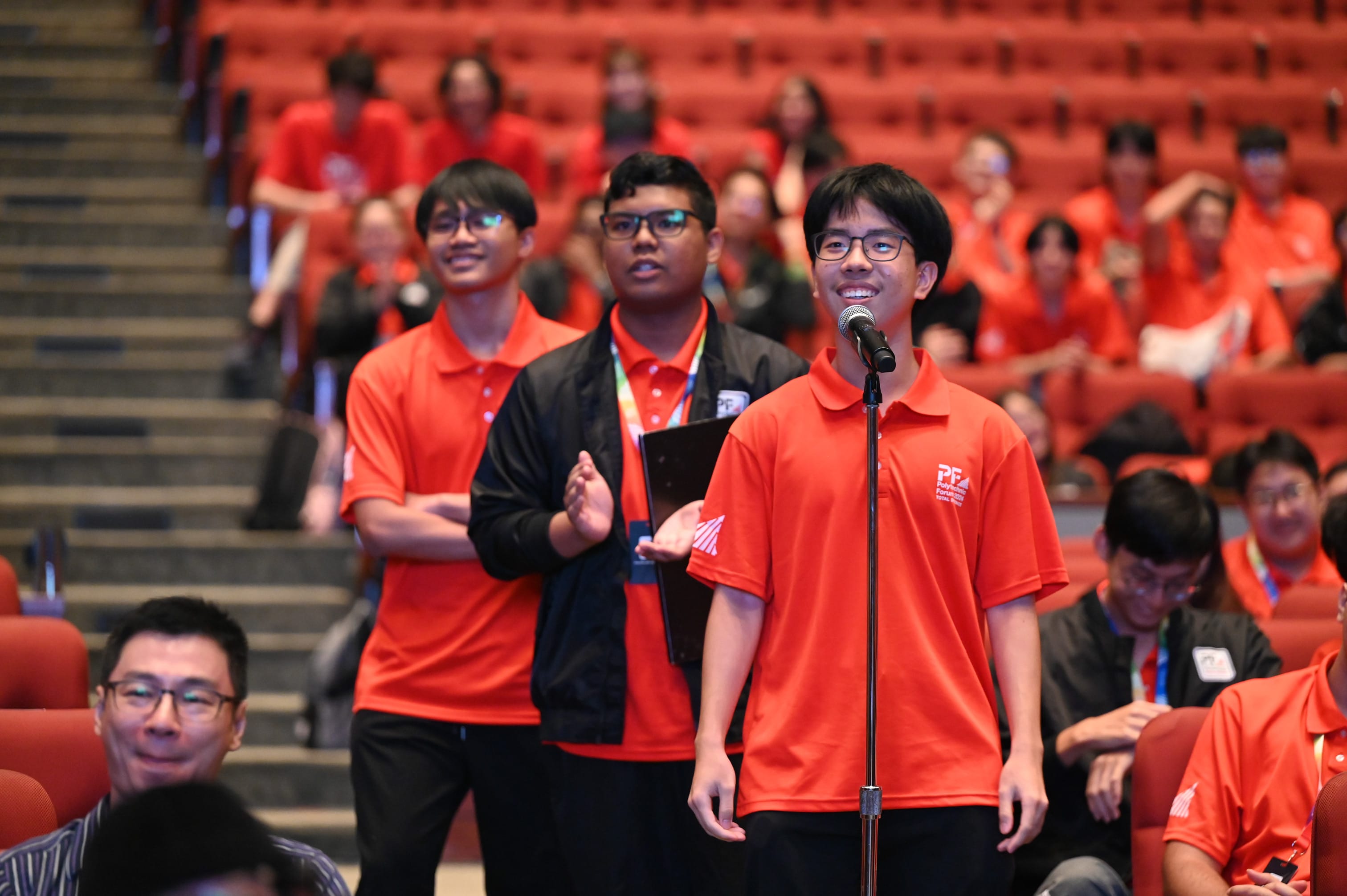 A young man in a red polo shirt speaks at a microphone in an auditorium filled with people in red shirts.