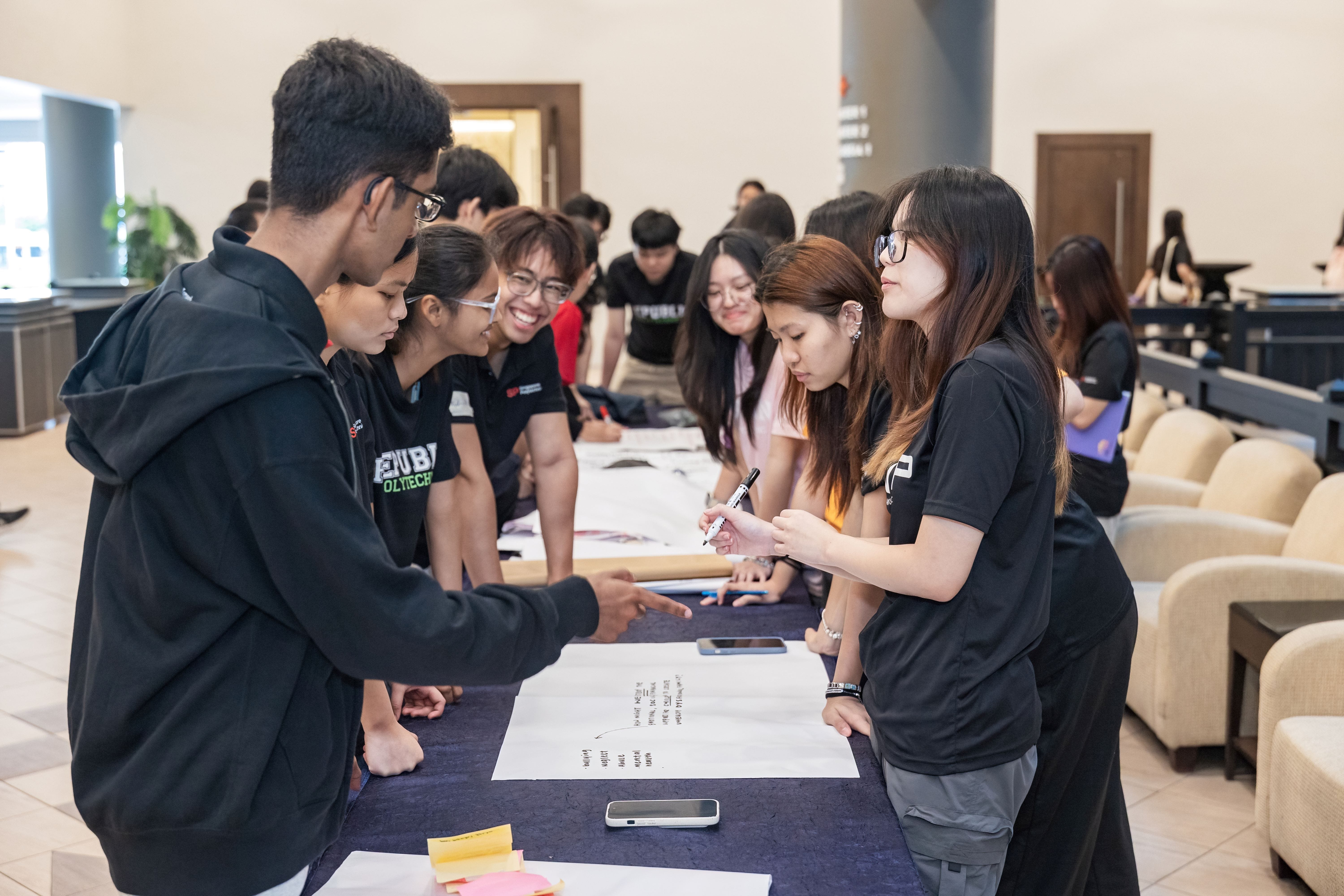 Students in black shirts stand around a table covered with large paper sheets, writing and discussing together indoors.