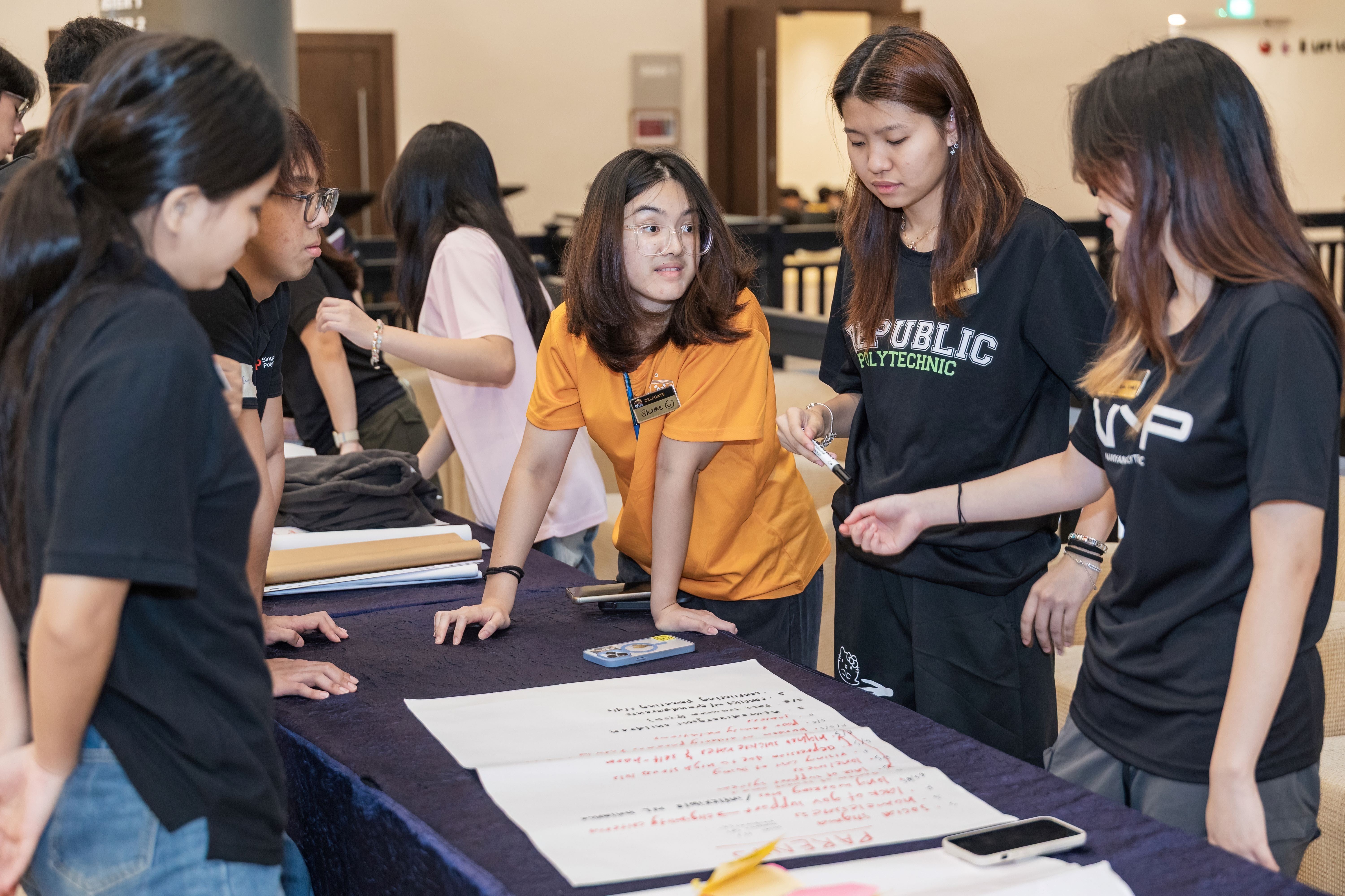 Students in black and orange shirts gather around a table with handwritten notes during a group activity.
