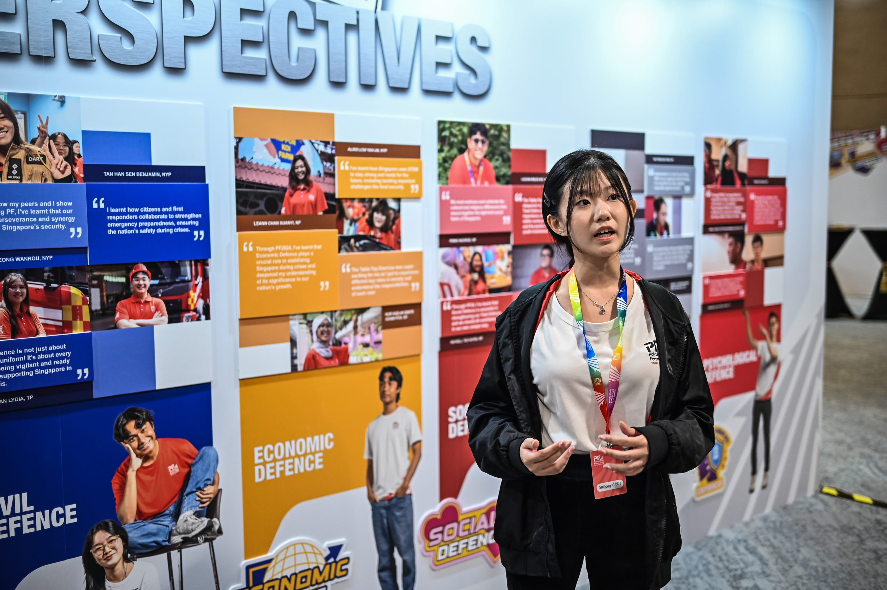 A young woman with a lanyard speaks in front of a display board titled Perspectives showing Civil and Economic Defence panels.
