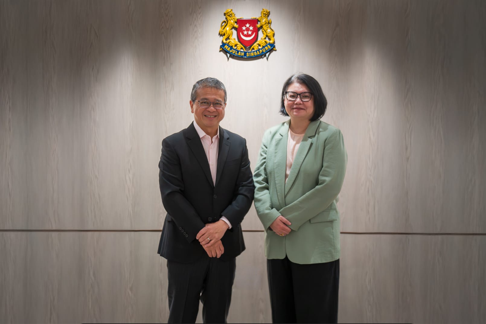 Two people stand smiling before the Singapore coat of arms on a wood-paneled wall.
