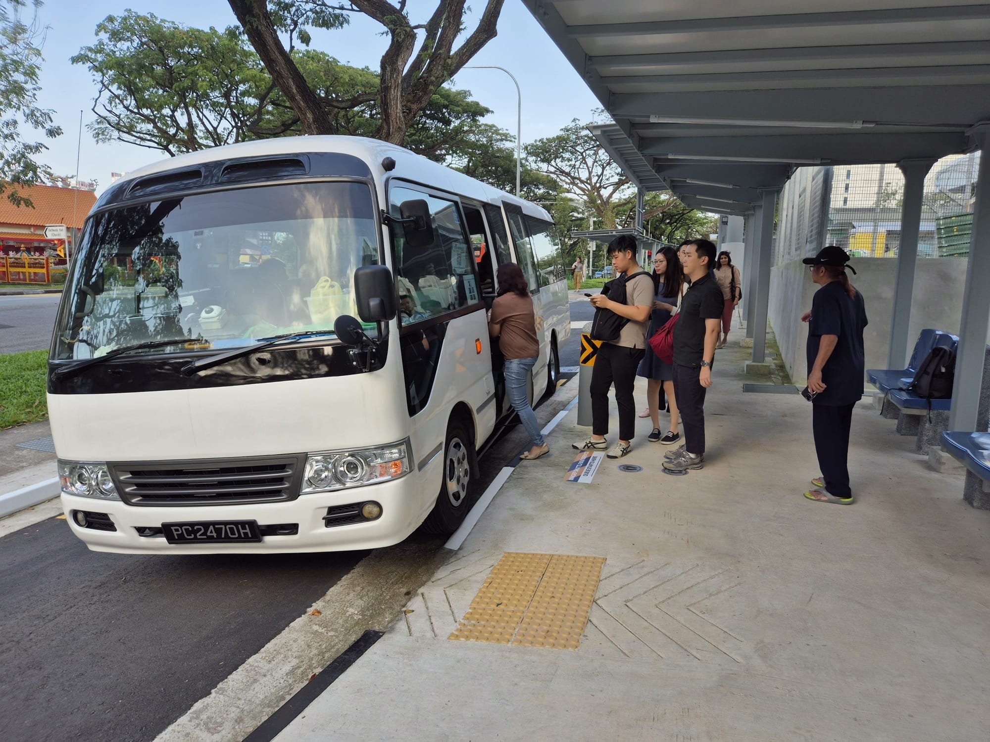 Several people queue to board a white minibus at a covered bus stop with blue seats and trees nearby.