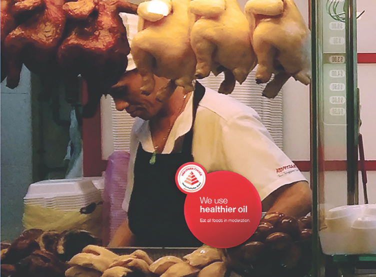 Food stall vendor preparing roasted meats with sign indicating use of healthier cooking oil.