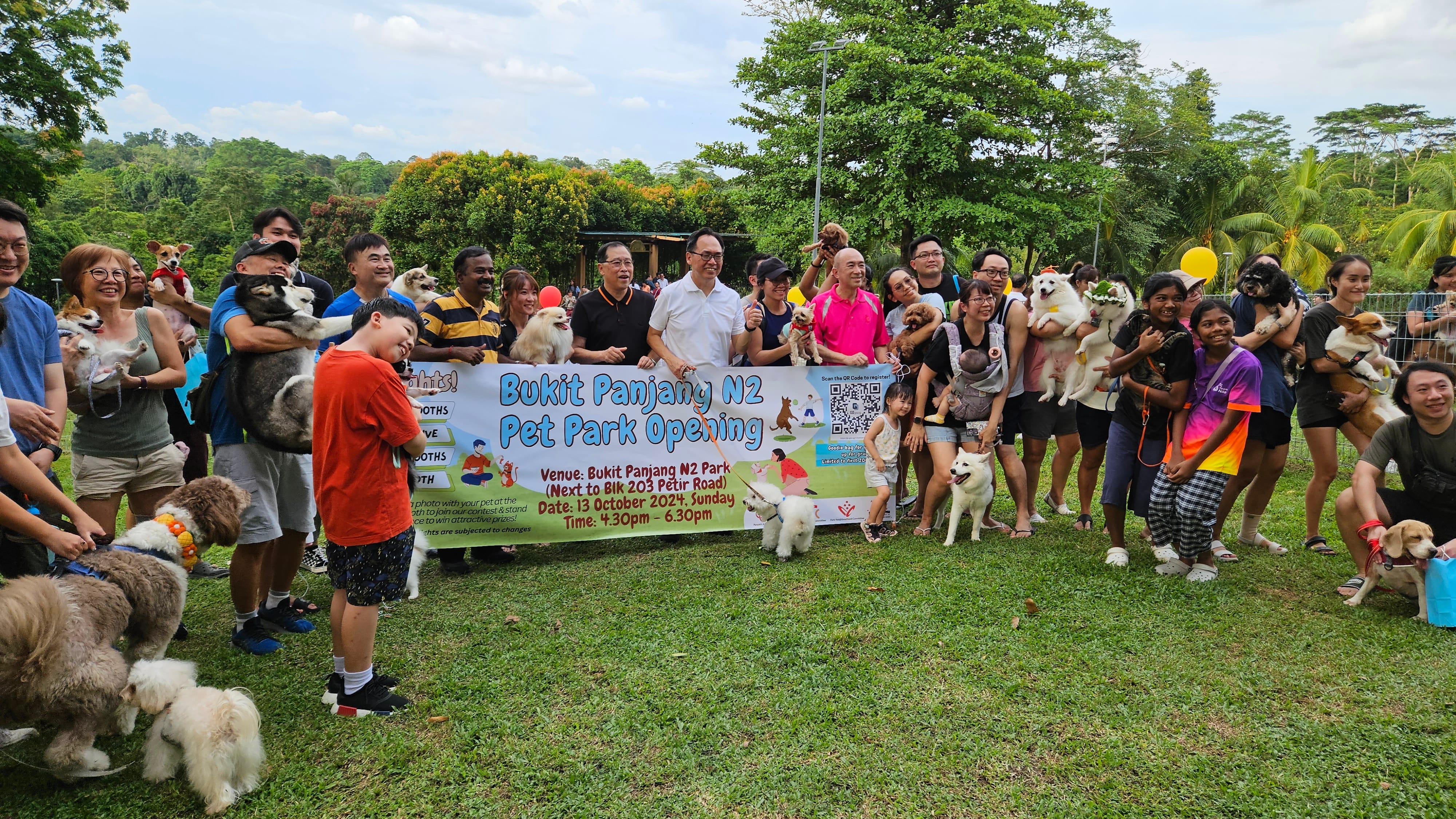 Group poses with dogs at "Bukit Panjang N2 Pet Park Opening" event on a grassy field.