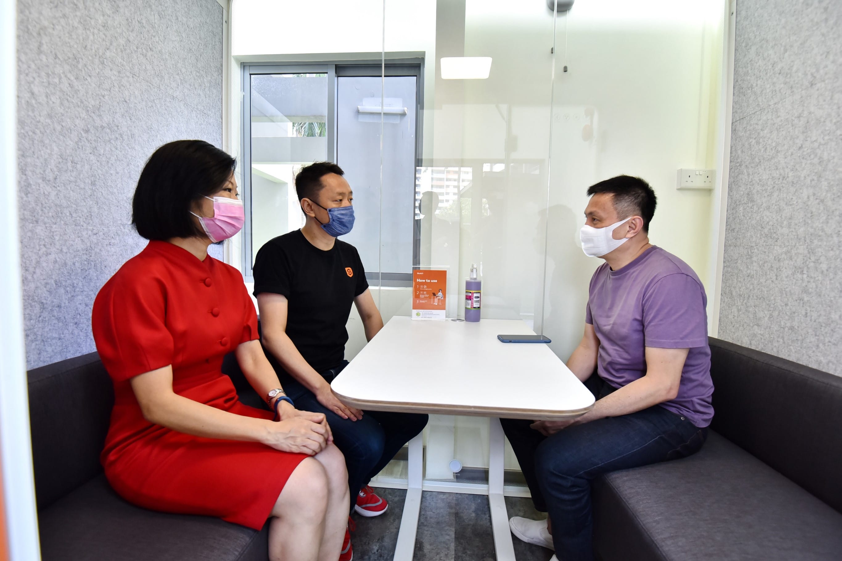 Three masked people sitting at a white table separated by a clear divider.