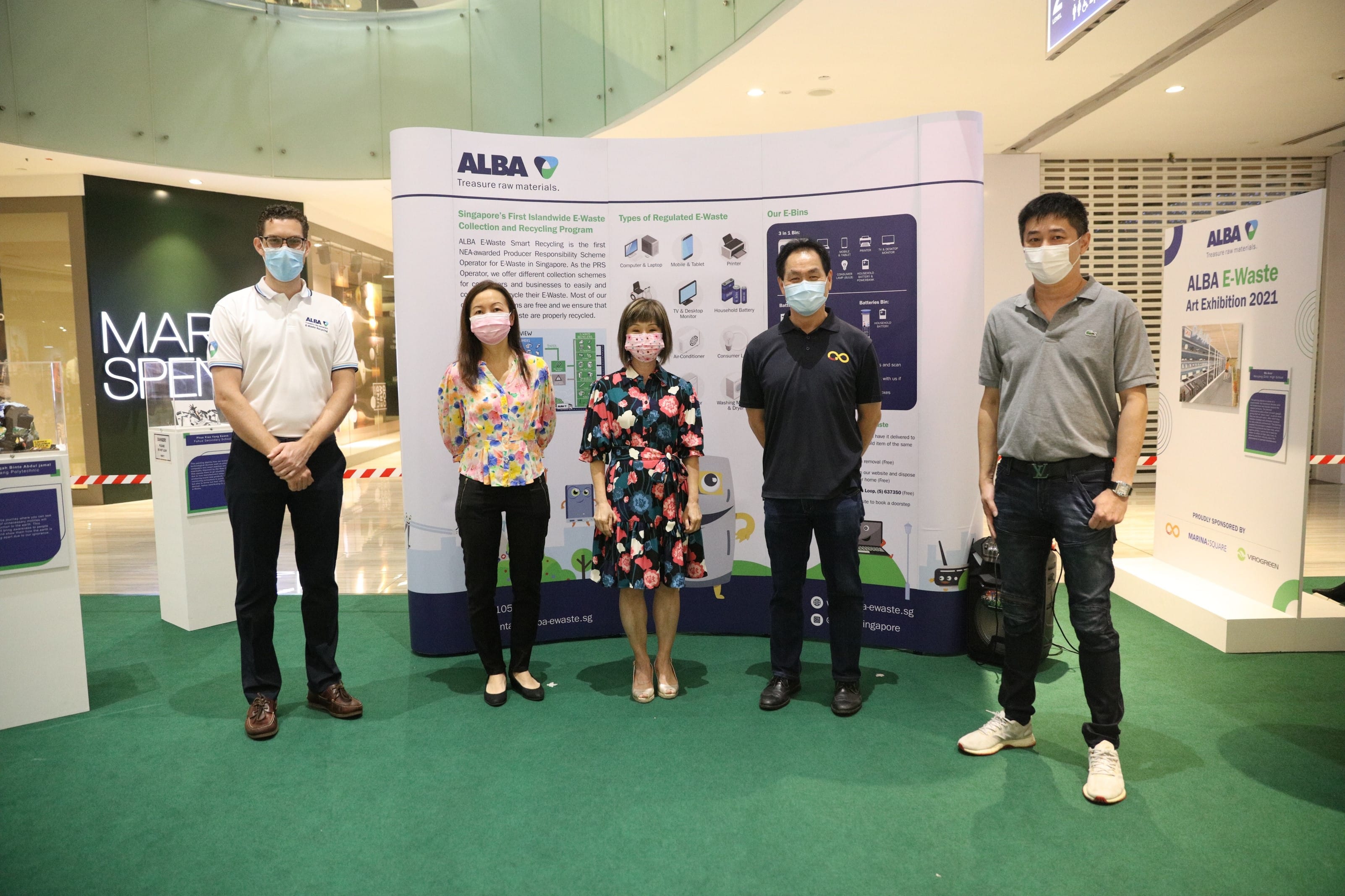 Five people in masks stand behind an "ALBA E-Waste Art Exhibition 2021" sign promoting recycling.