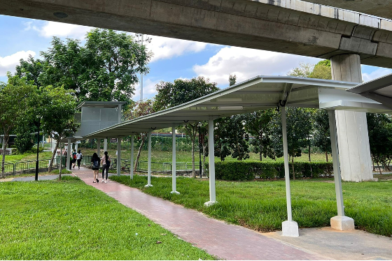 Covered walkway on a brick path, grass on either side, beneath a concrete structure. People walking.
