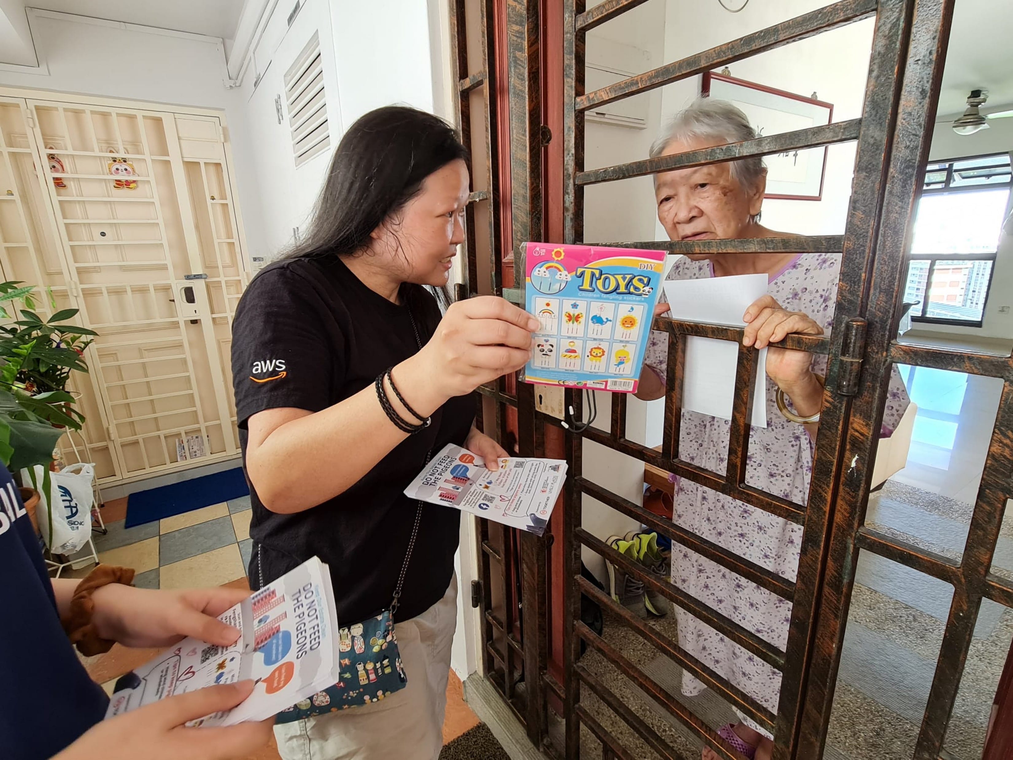 Woman in AWS shirt shows "Toys" sticker pack to senior woman through gate. Another flyer visible.