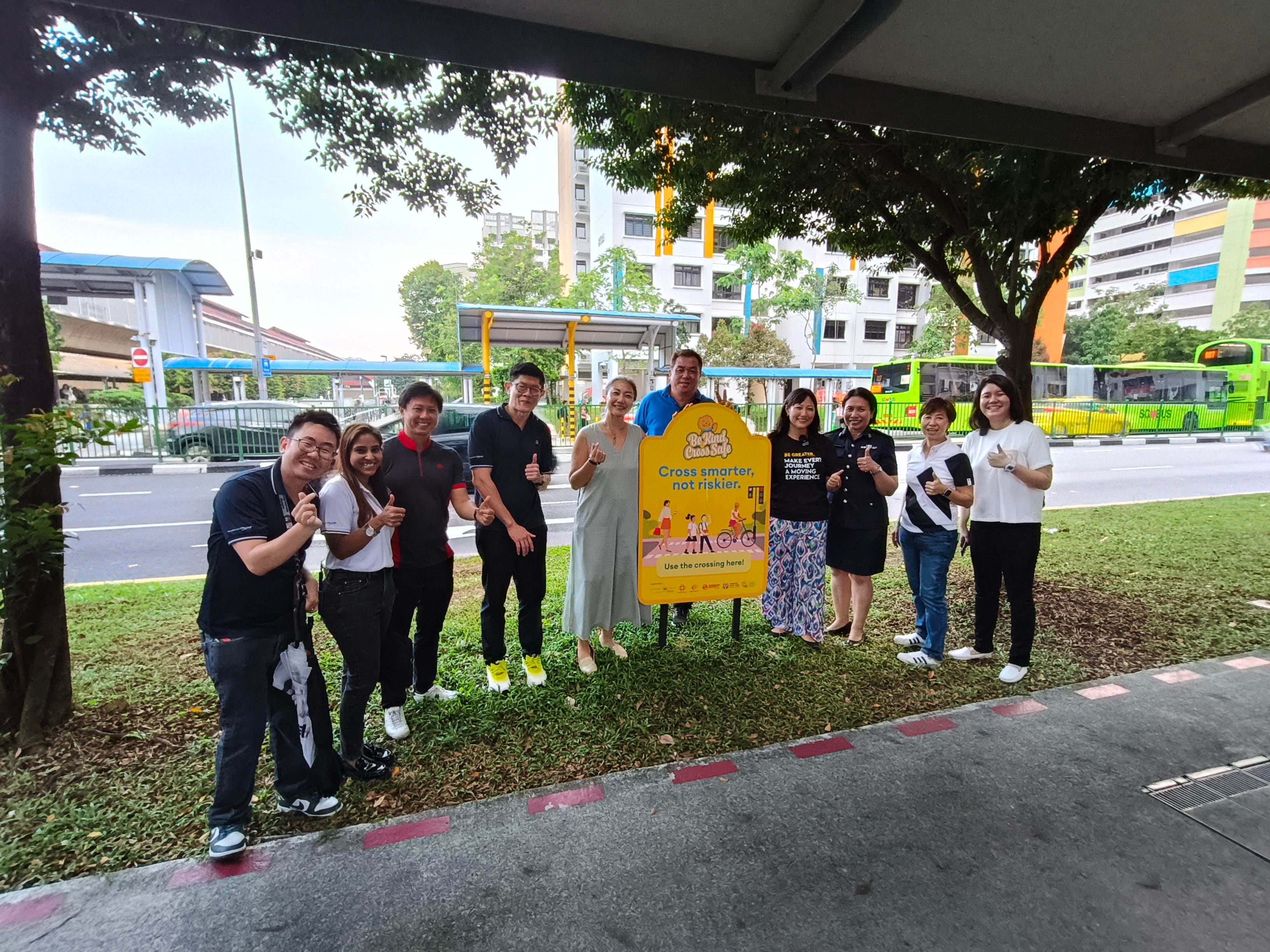 Group of people pose by "Cross smarter, not riskier" safety sign near a road with a green SBS bus.
