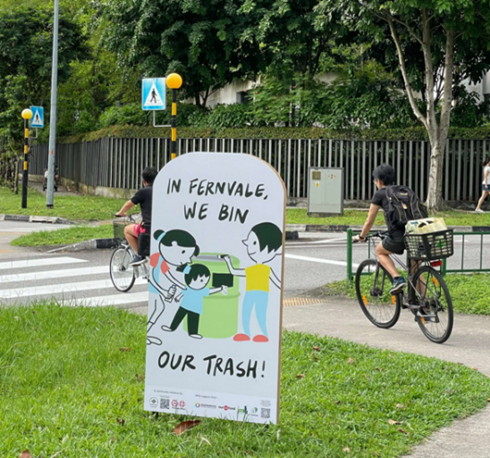“In Fernvale, we bin our trash!” sign beside cyclists at crossing.