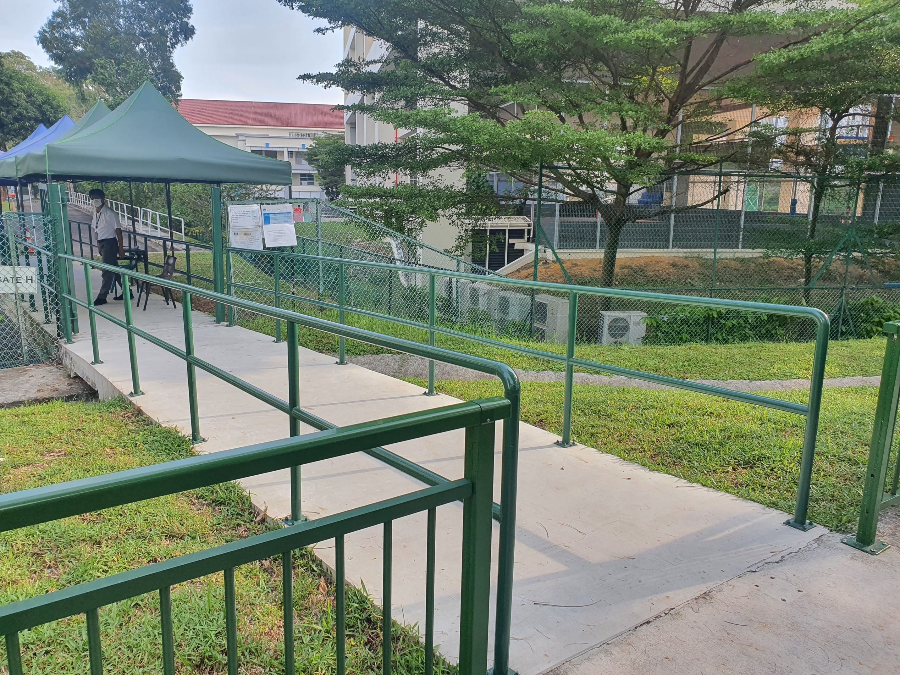 Green railings and a paved ramp leading to a tented area; a person stands in the background.
