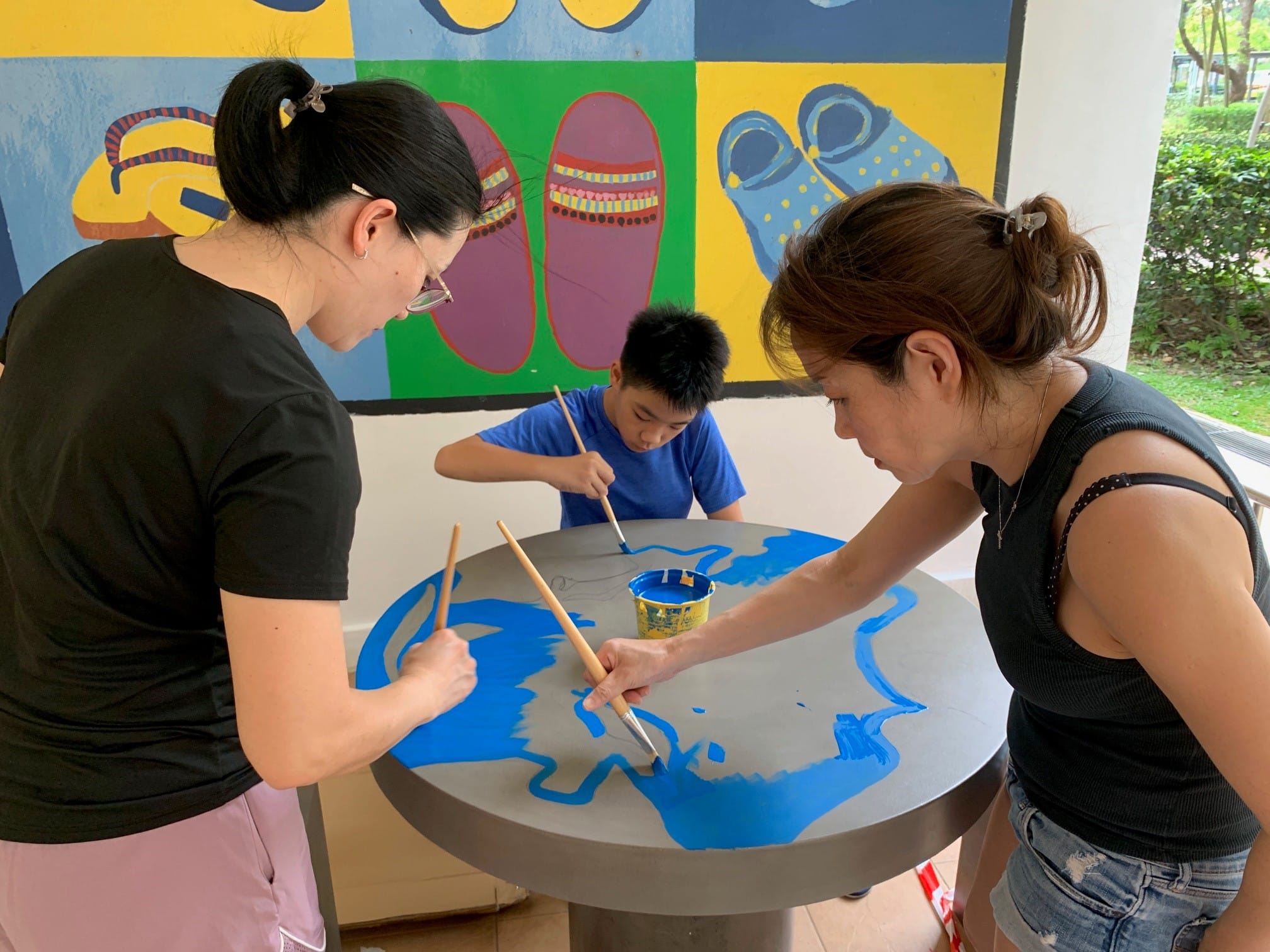 Two adults and child painting a world map in blue on a circular gray table. Mural of sandals behind.
