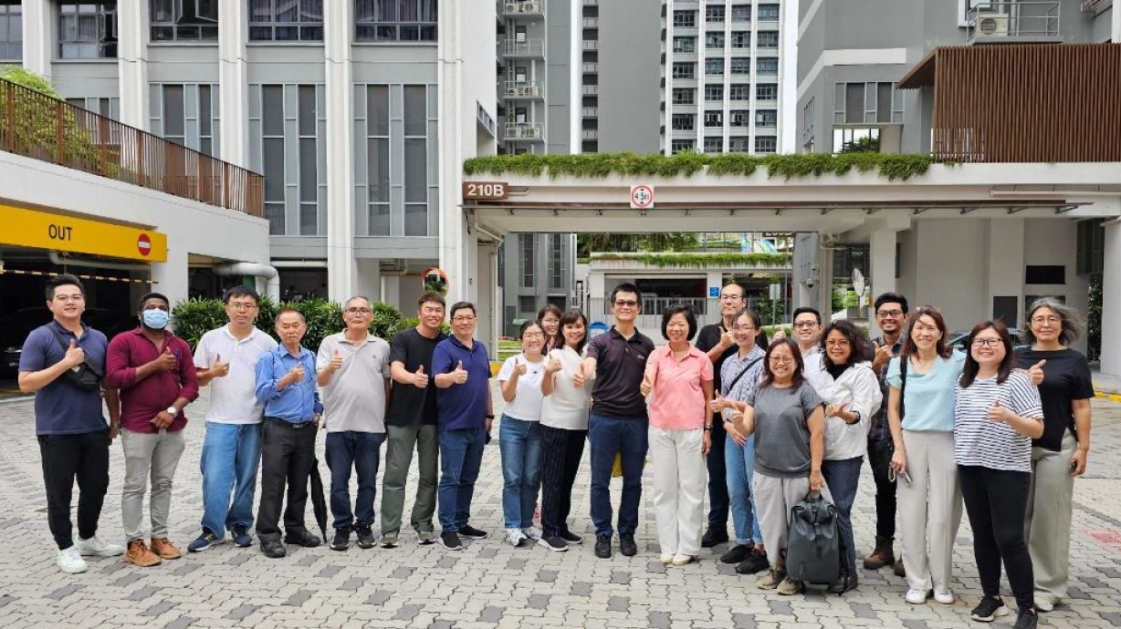 Group of about 20 people give thumbs-up outside a building with "210B" sign.