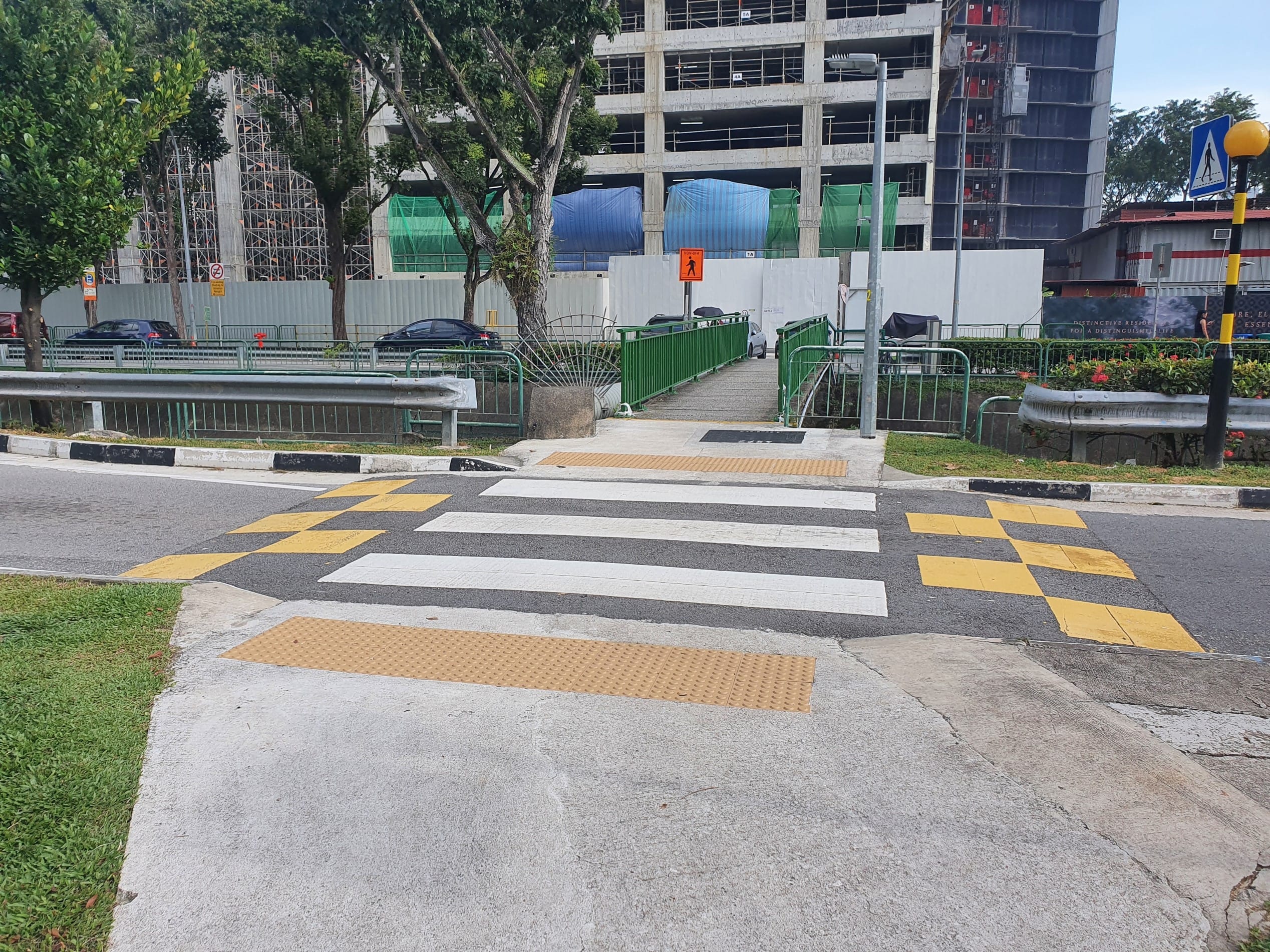 Crosswalk with yellow tactile paving, leading to a green pedestrian bridge near a construction site.