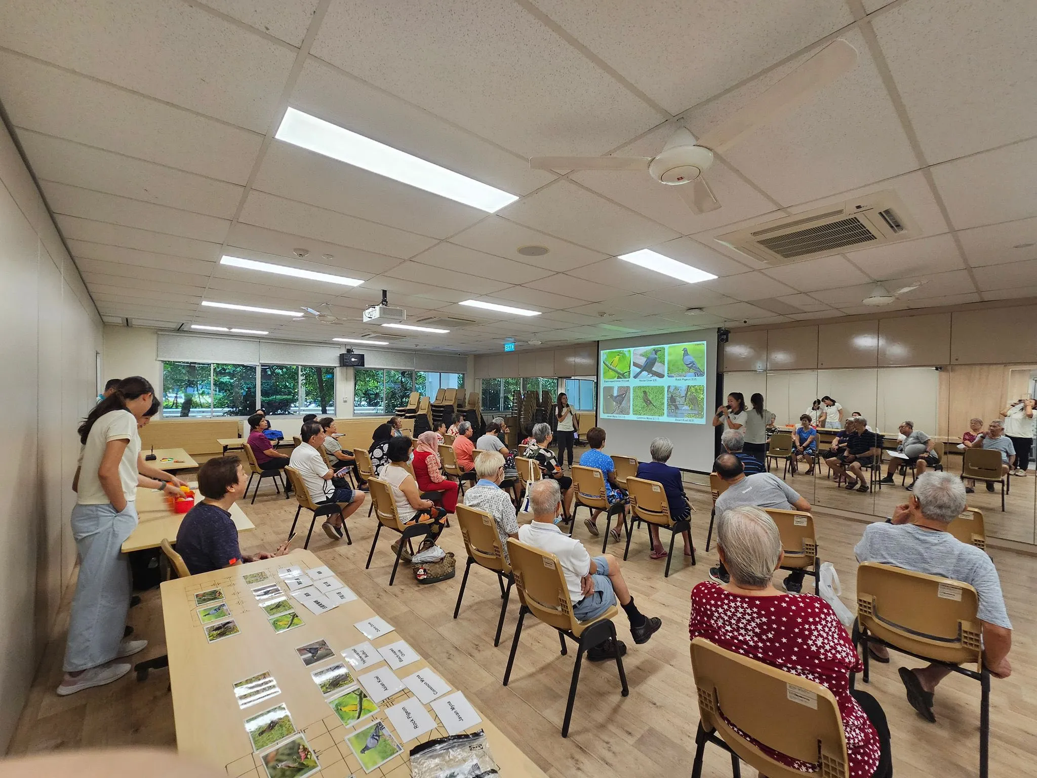 Indoor classroom with people seated for presentation on birds, cards on tables.