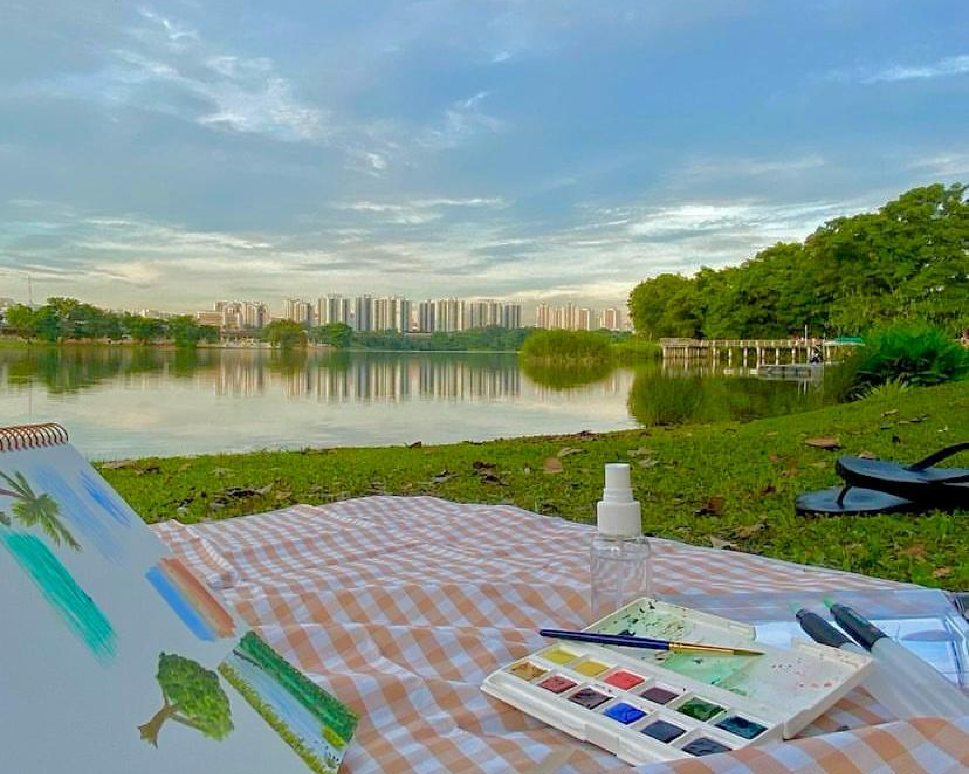 Watercolor paints and sketchbook on a checkered blanket beside a calm lake with city buildings in the background.