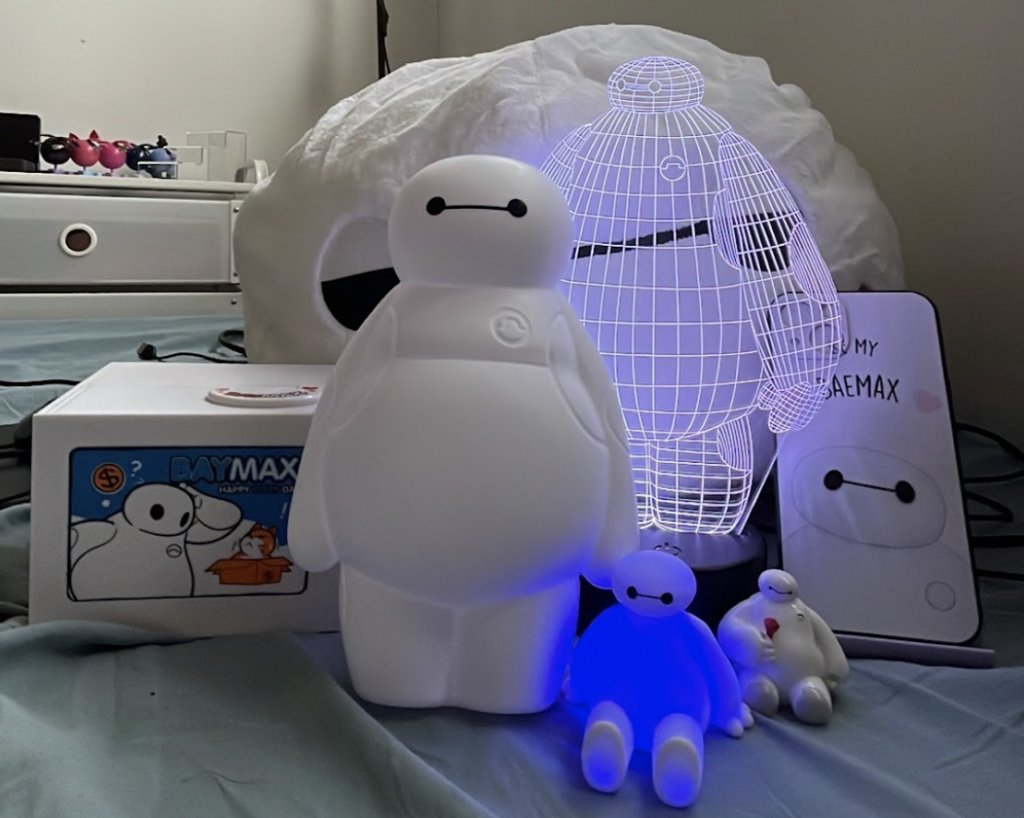 Multiple Baymax figures including a lamp, glowing 3D wire LED light, small plush, and a Baymax box on a blue surface.