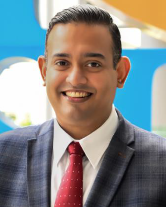 Man in a gray blazer and red tie smiling, with a colorful blue and yellow background.
