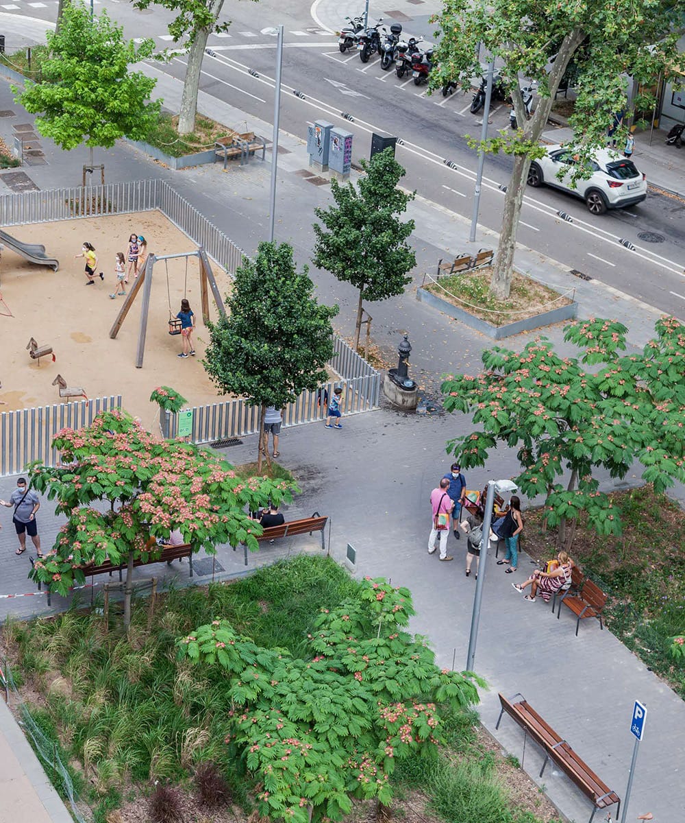 High-angle, overhead shot of a lively urban plaza and pedestrian park in Barcelona. On the left, children play inside a fenced, sandy playground equipped with swings and a slide. Lush green trees with vibrant pink blossoms and manicured grass patches line a stone-paved walkway where pedestrians gather near a small food cart and sit on wooden benches. A traditional dark cast-iron public drinking fountain stands in the center of the path. In the background, a white car drives down a multi-lane city street flanked by parked motorcycles and roadside trees.