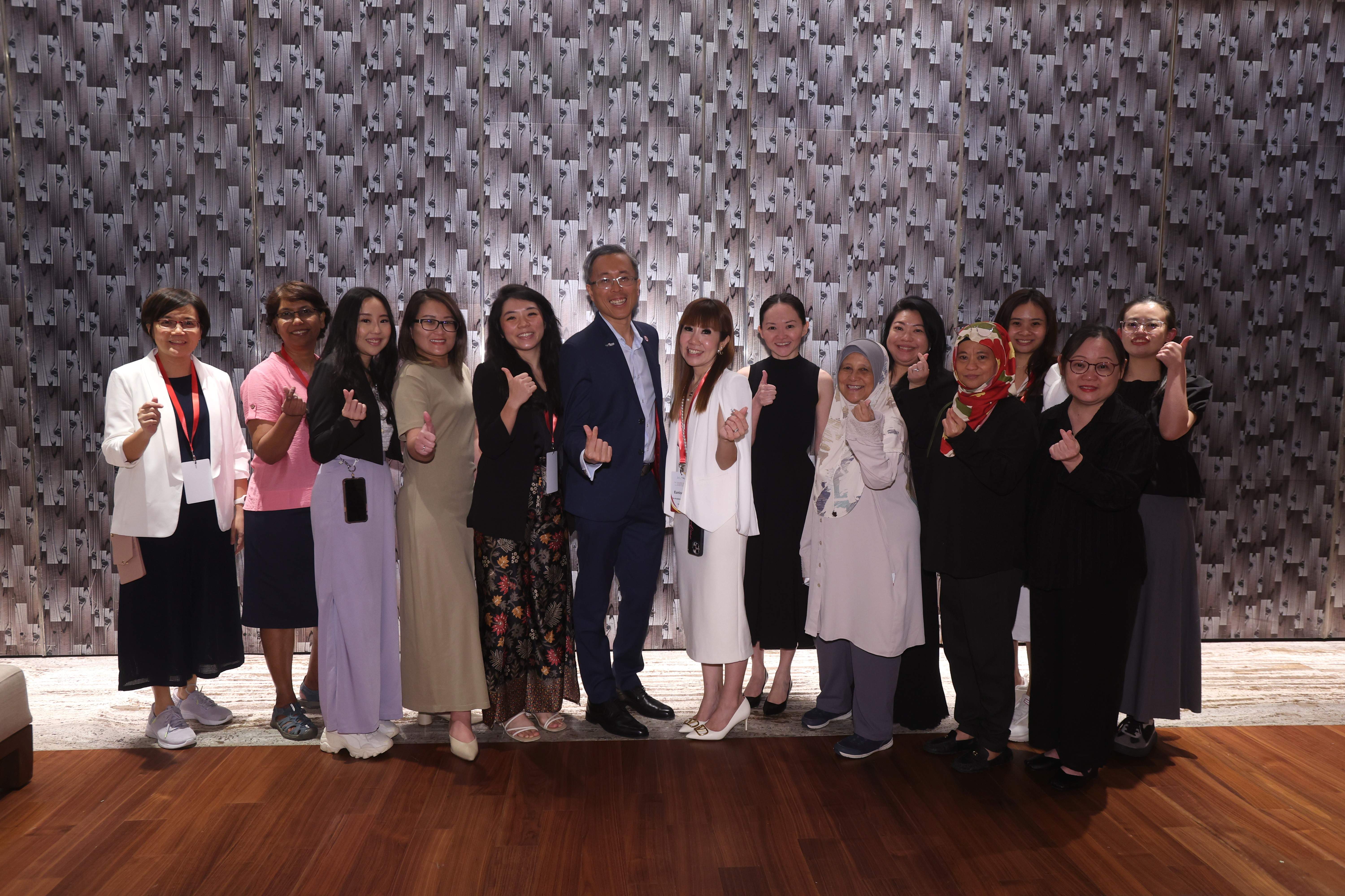 A group of about 14 people posing together indoors, making thumbs-up and finger-heart gestures, in front of a patterned wall.