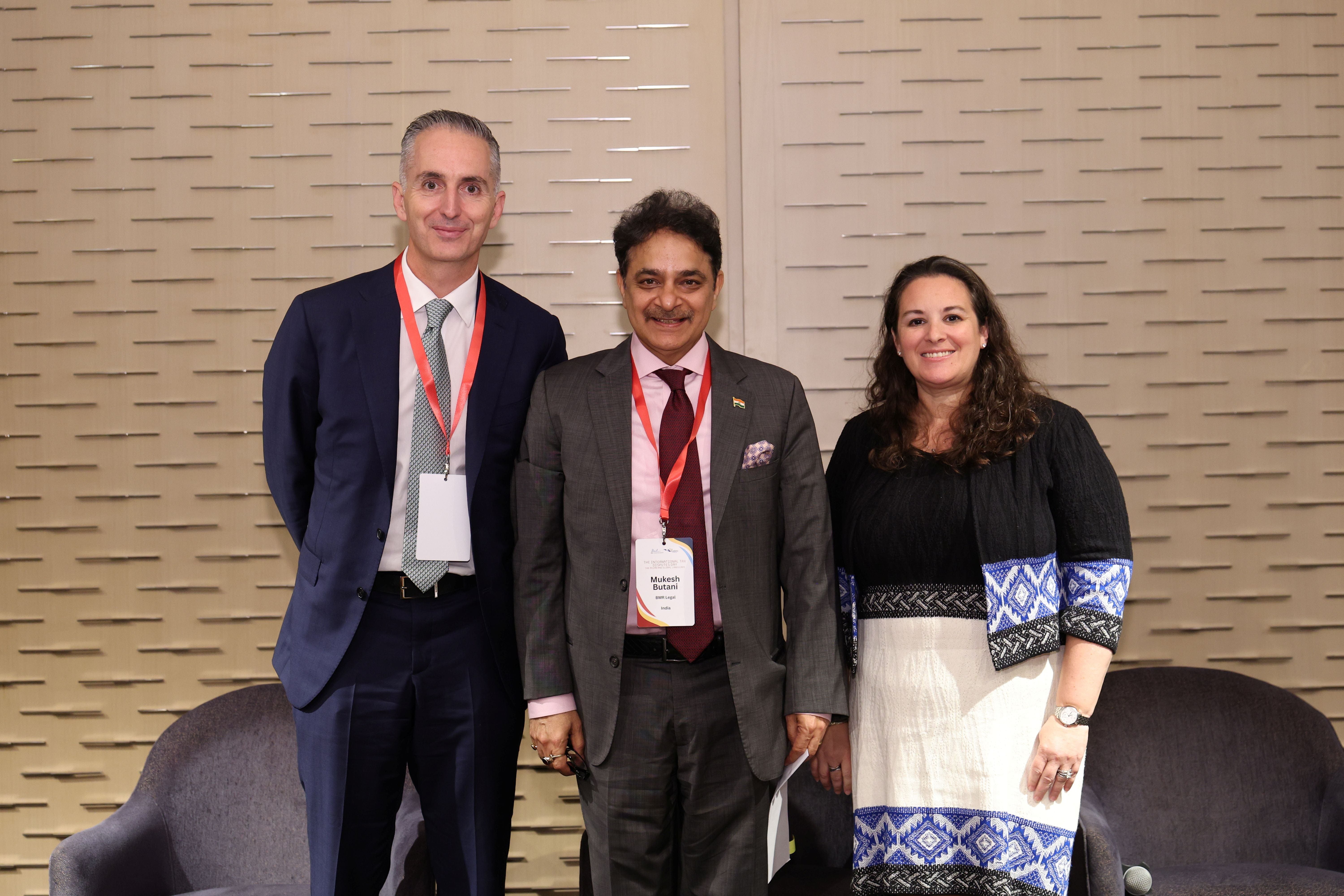 Three smiling conference attendees wearing name badge lanyards pose together in front of a textured beige wall.