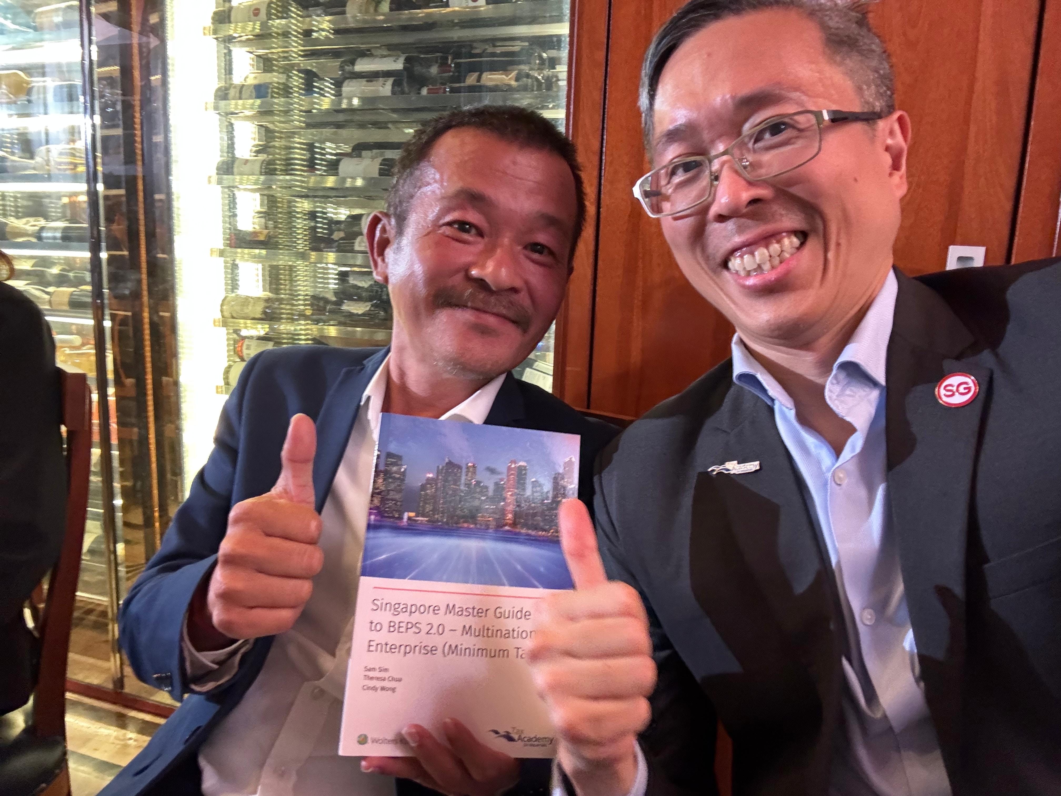 Two smiling men give thumbs up while holding a book titled Singapore Master Guide to BEPS 2.0 Multinational Enterprise.