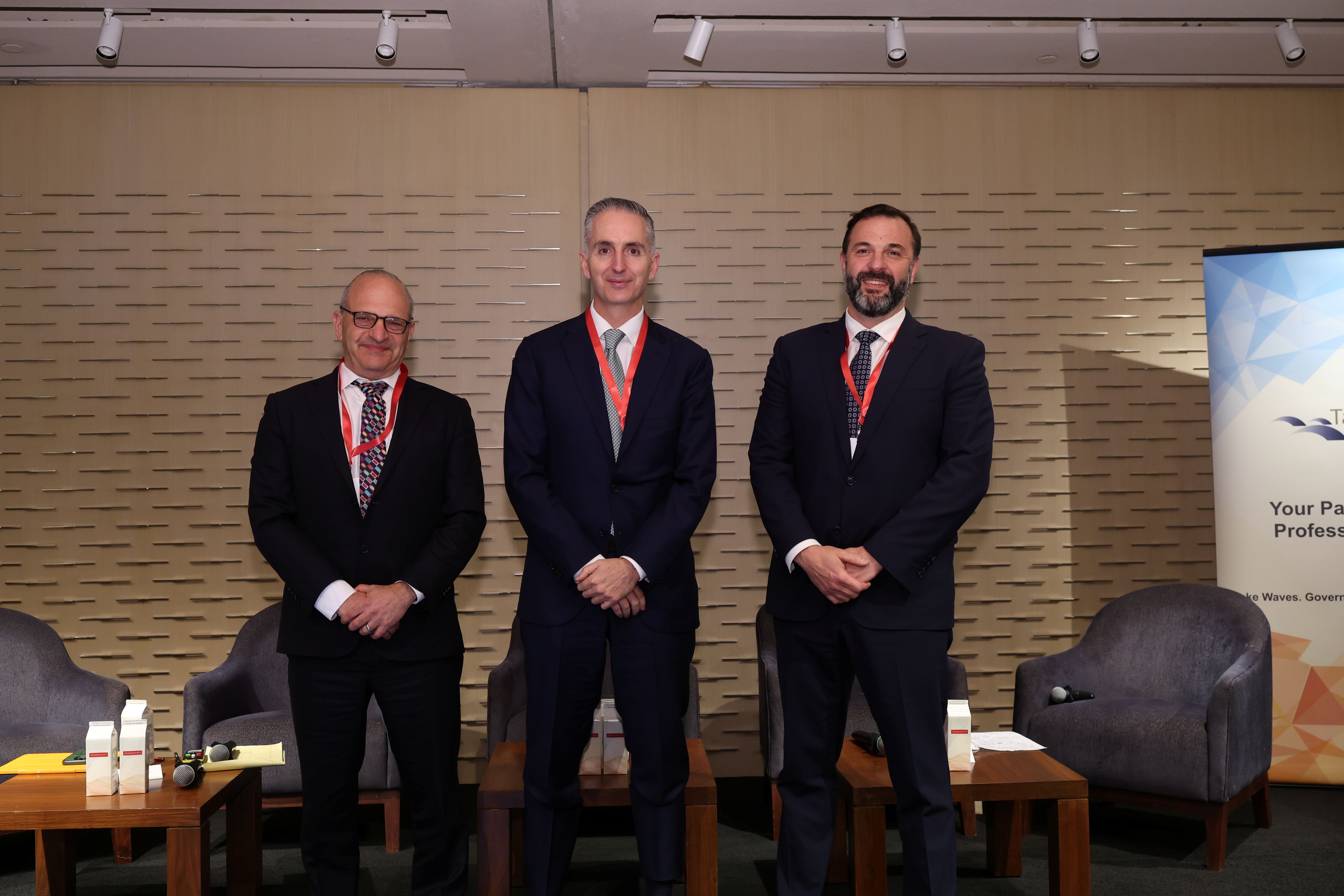 Three men in dark suits with red lanyards stand together at a professional conference event.
