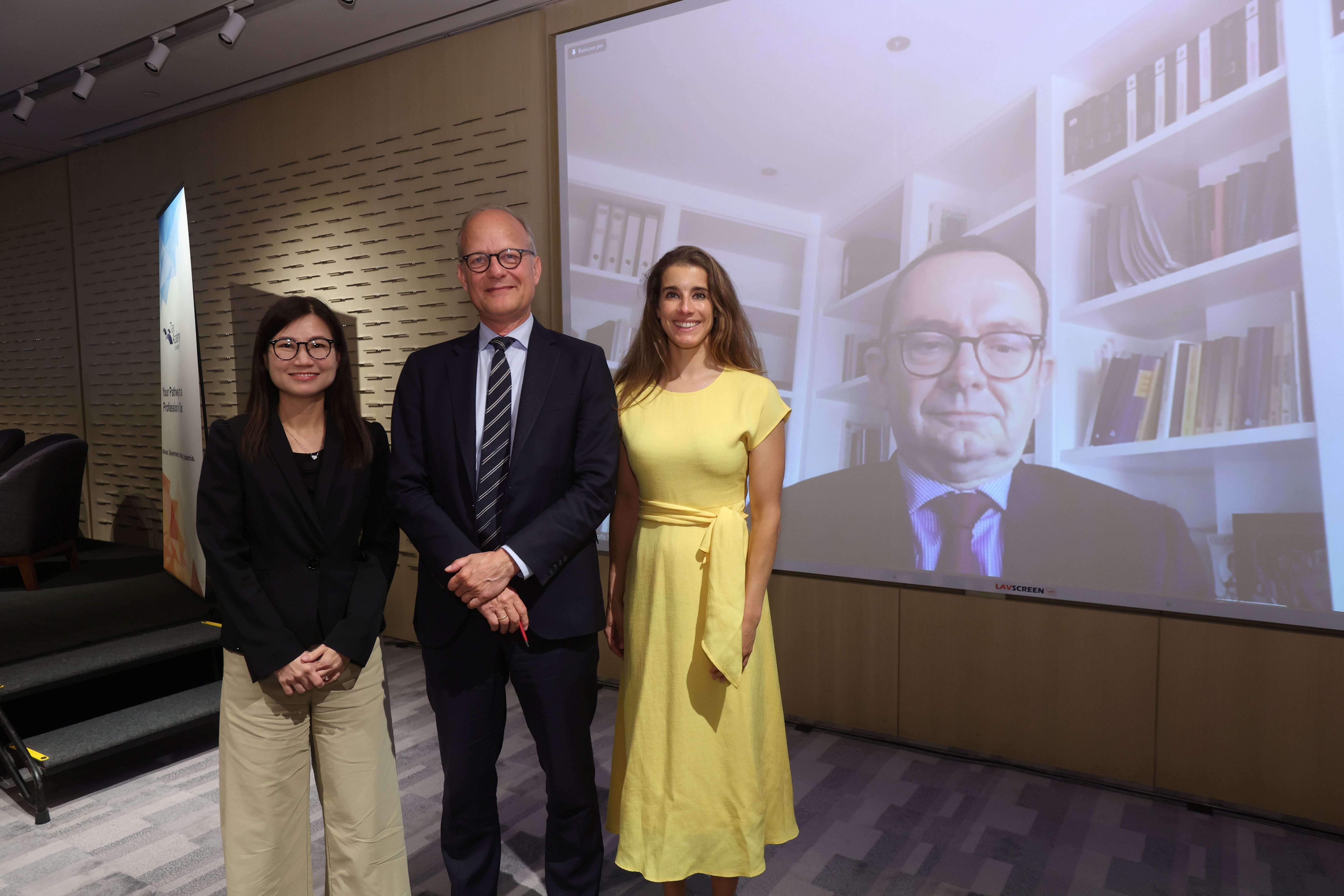 Three people stand in a conference room posing beside a large screen showing a man on a video call.