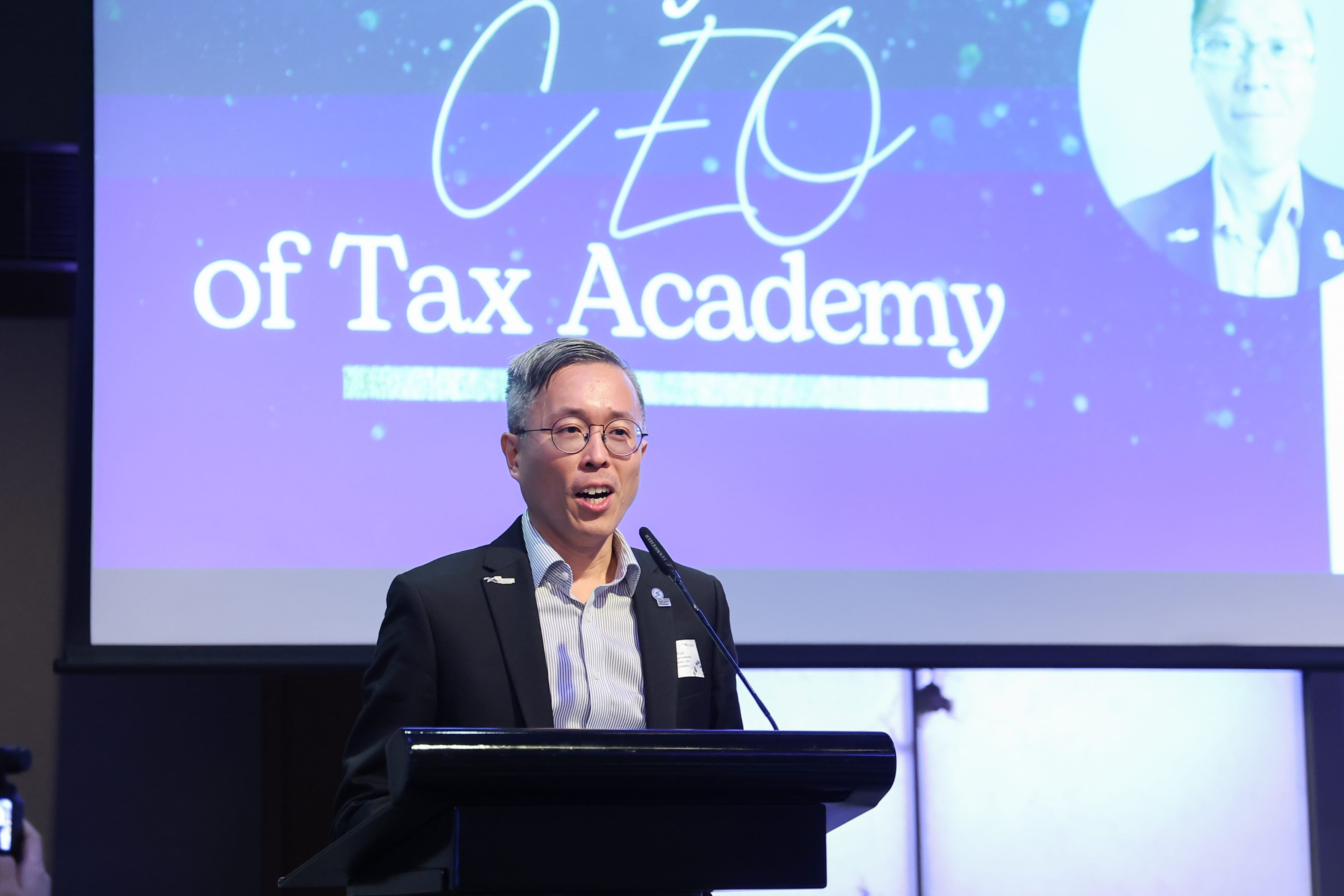 A man with glasses speaks at a podium in front of a large screen reading "CEO of Tax Academy."