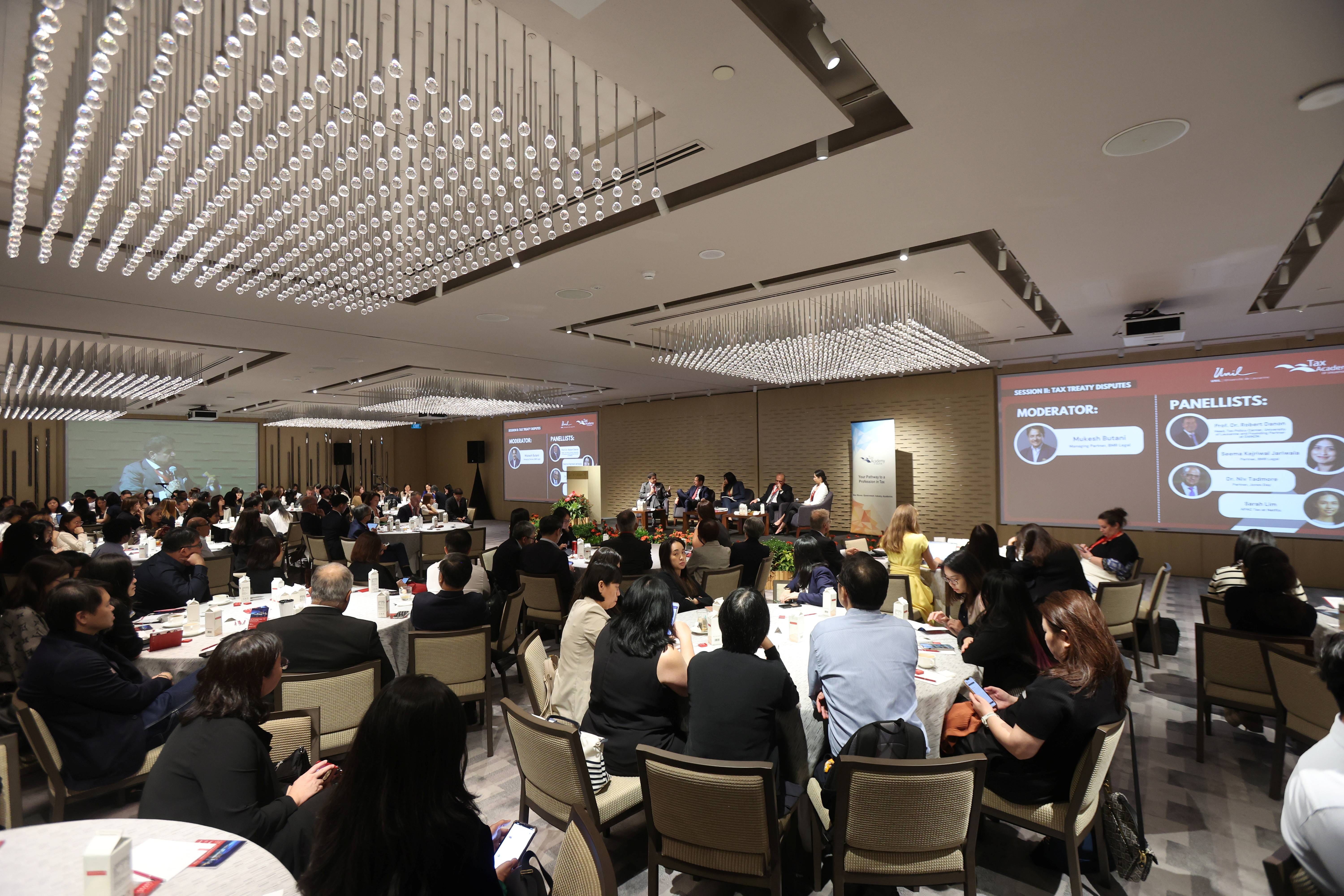 A large conference room filled with attendees at round tables, with panelists on stage and presentation screens showing moderator and panellists.