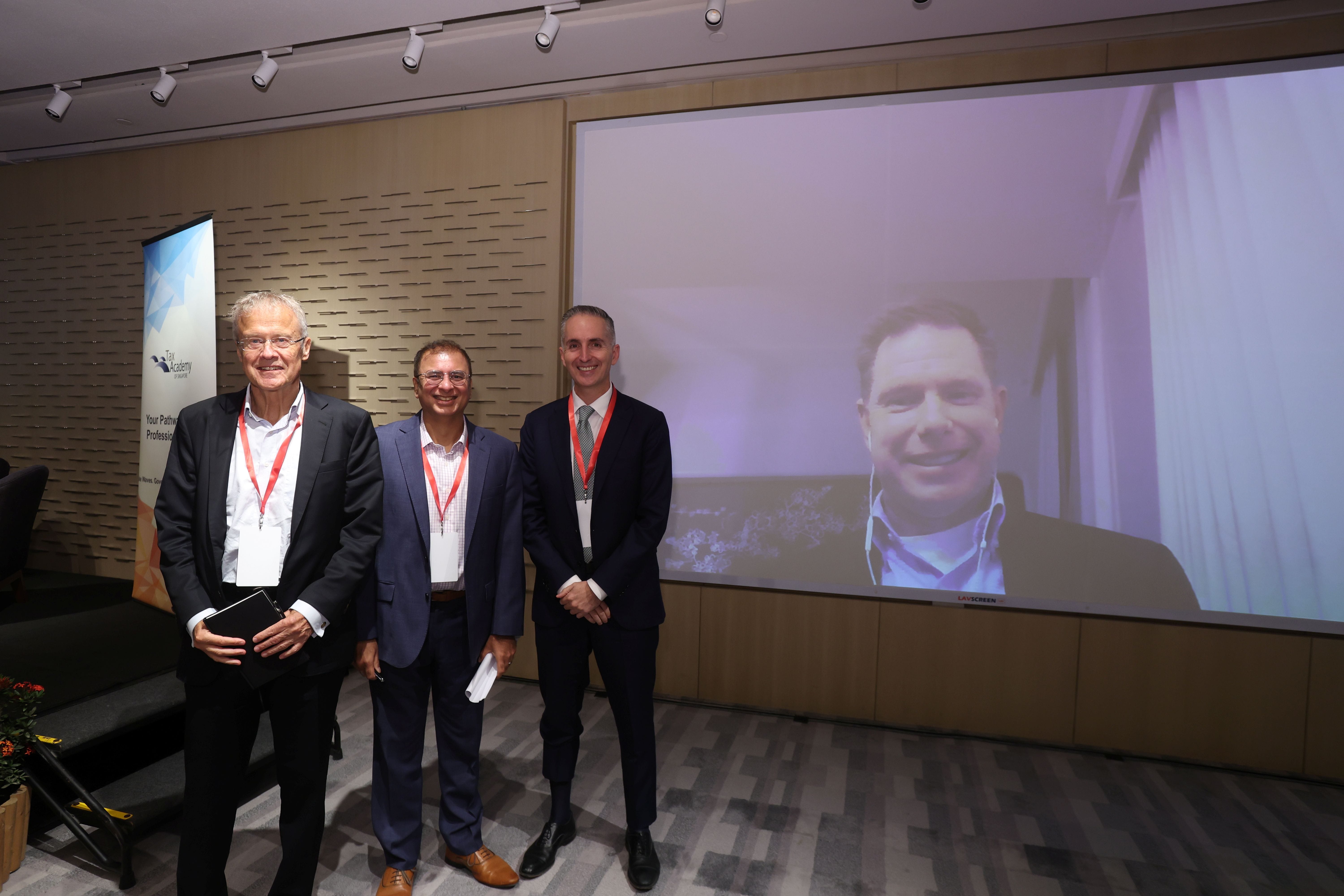 Three men in suits with red lanyards stand before a large screen showing a fourth man joining via video call.