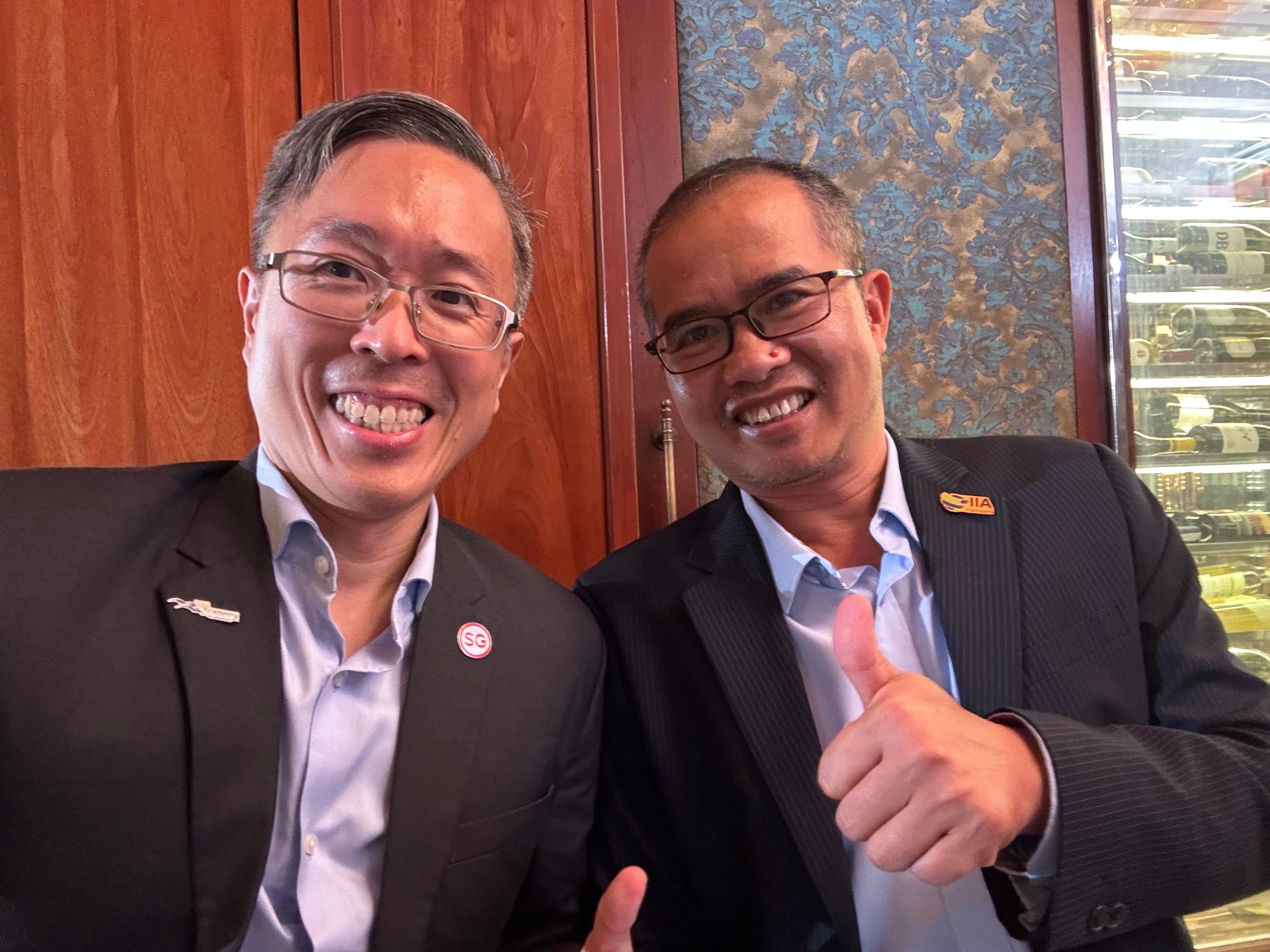 Two smiling men in dark suits with lapel pins giving thumbs up, seated near a wood panel and wine rack.