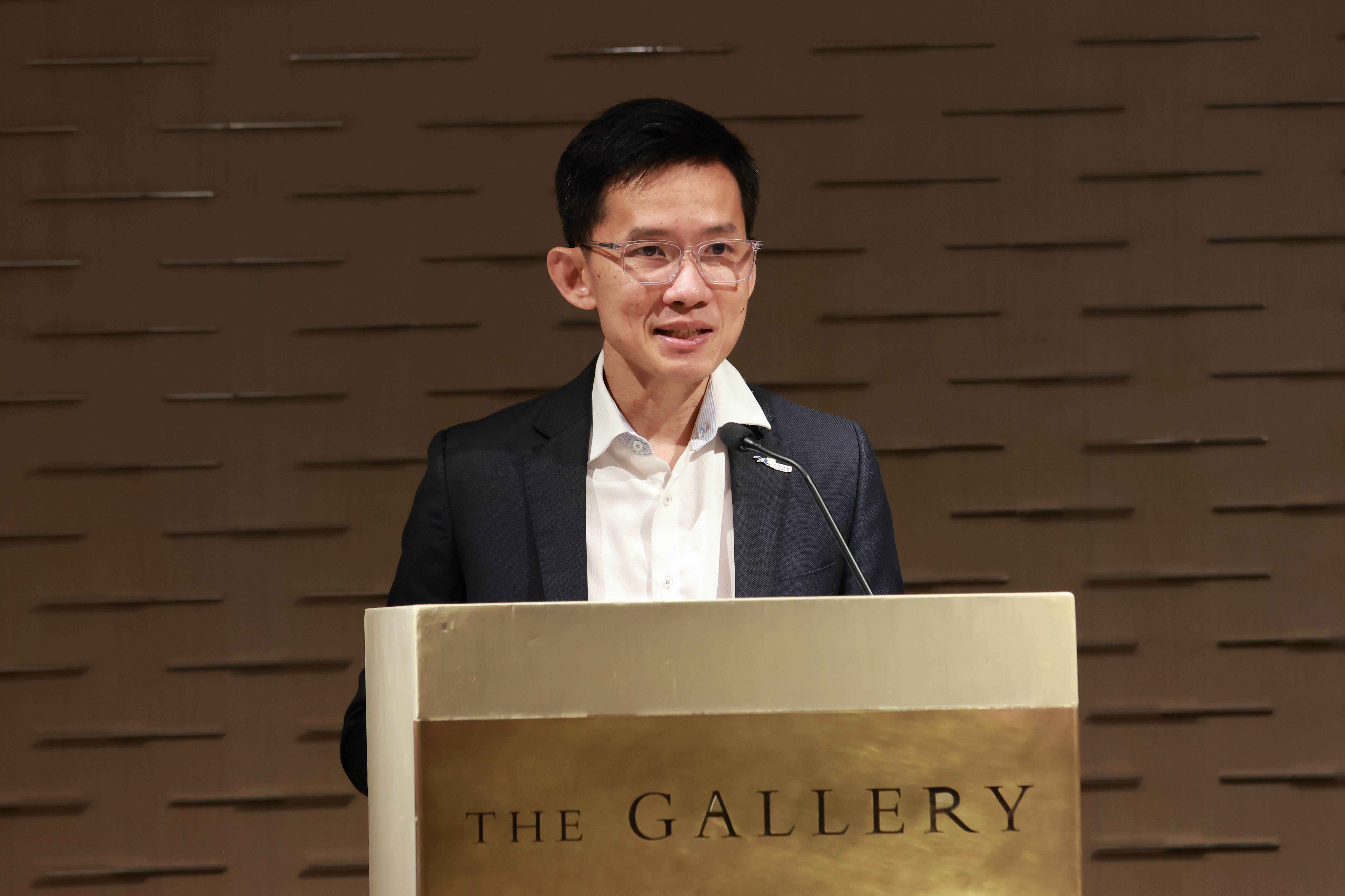 A man wearing glasses and a dark blazer speaks at a podium labeled "The Gallery" with a microphone.