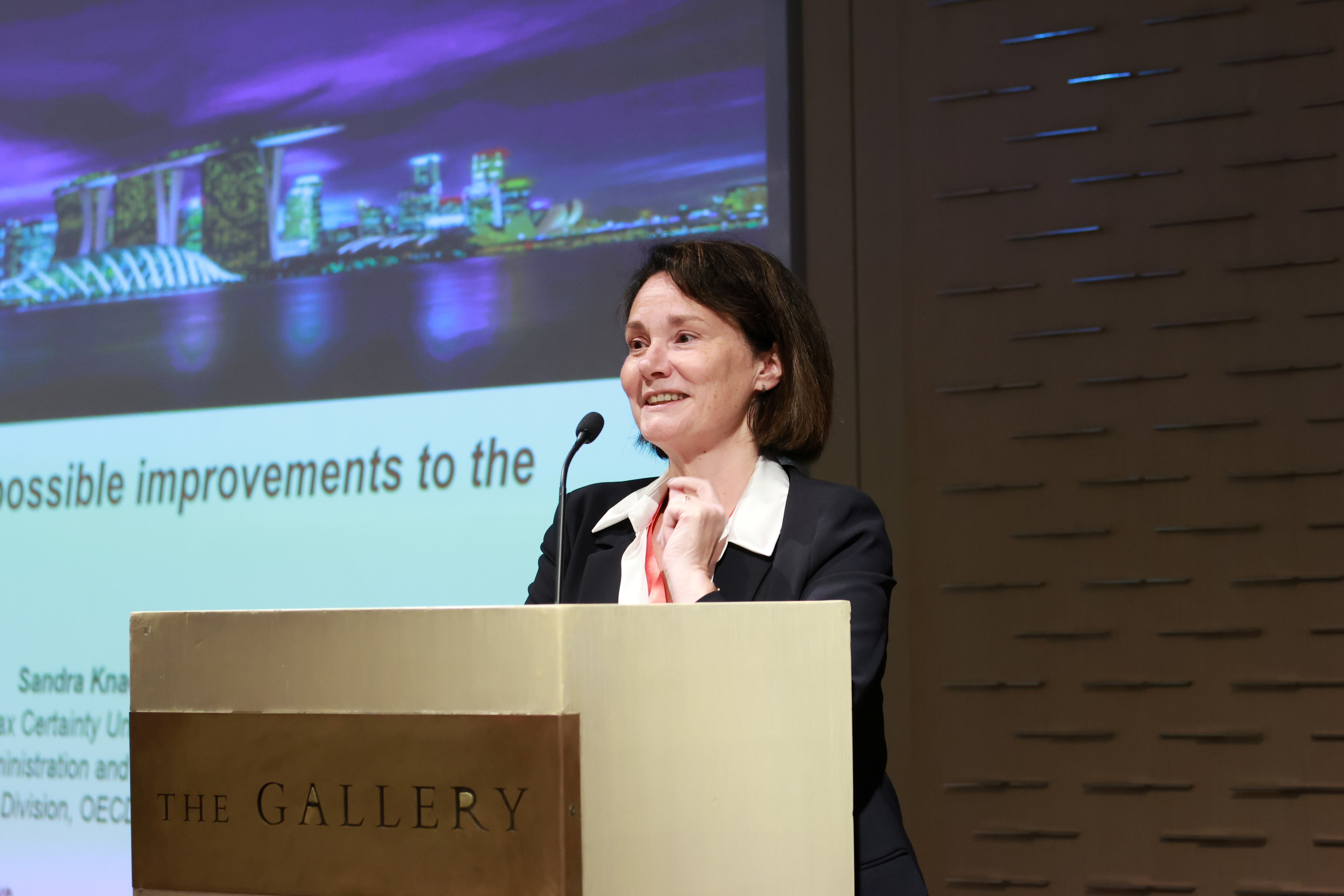 Woman in a dark blazer speaking at a podium labeled "The Gallery" with a presentation slide visible behind her.