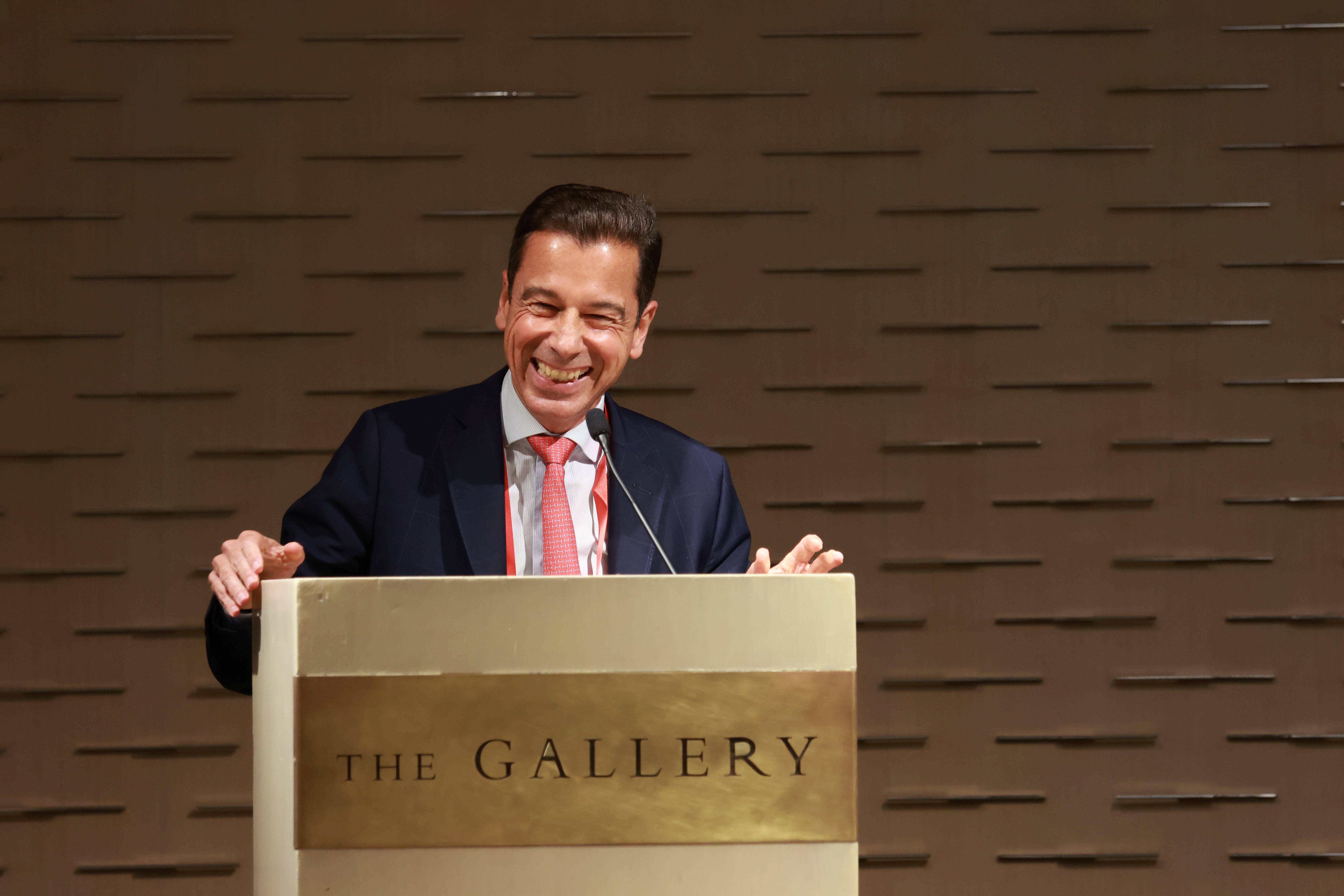 A smiling man in a navy suit and red tie speaks at a podium labeled "The Gallery."