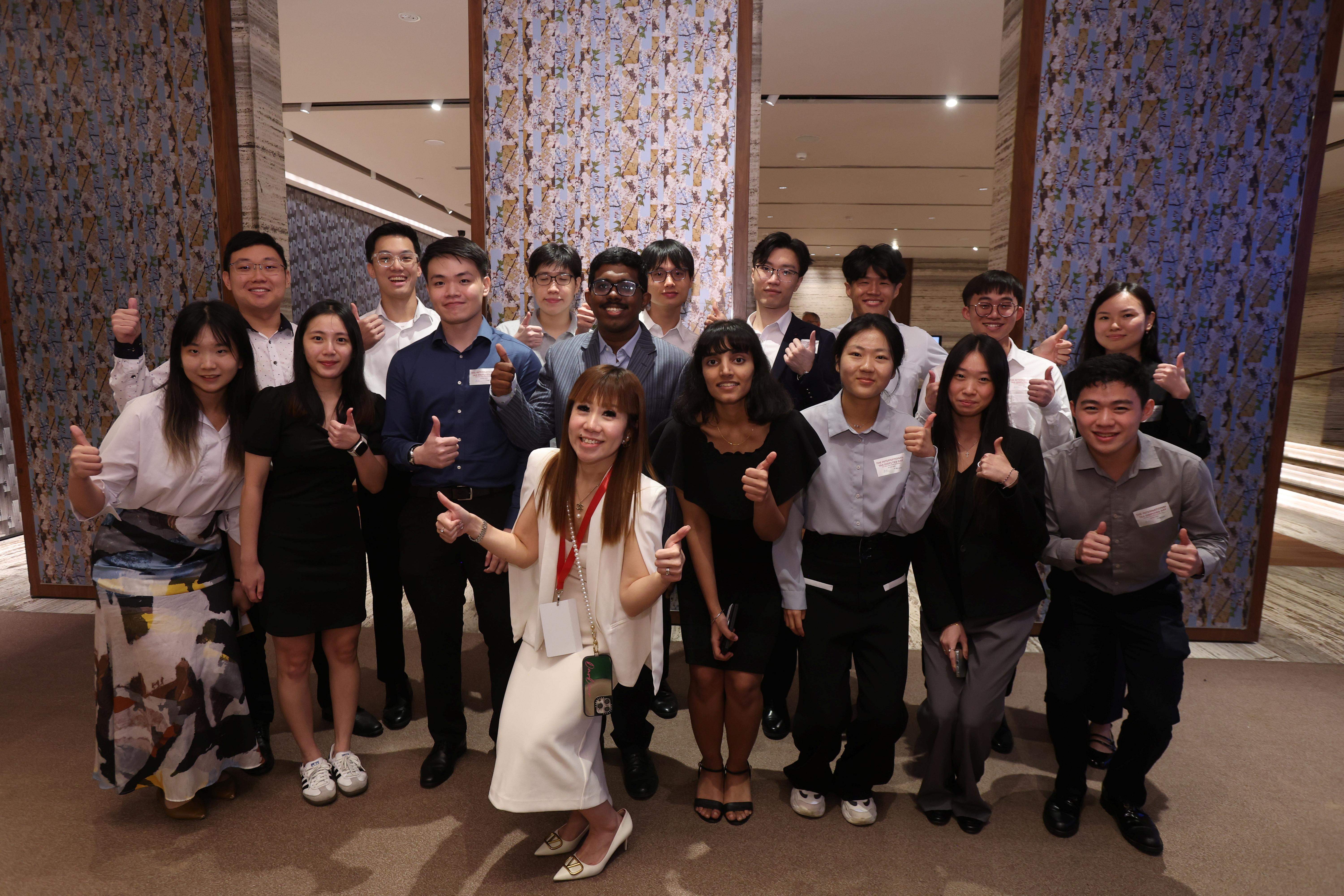 A group of about 20 young adults posing with thumbs up indoors in front of a decorative tiled wall.