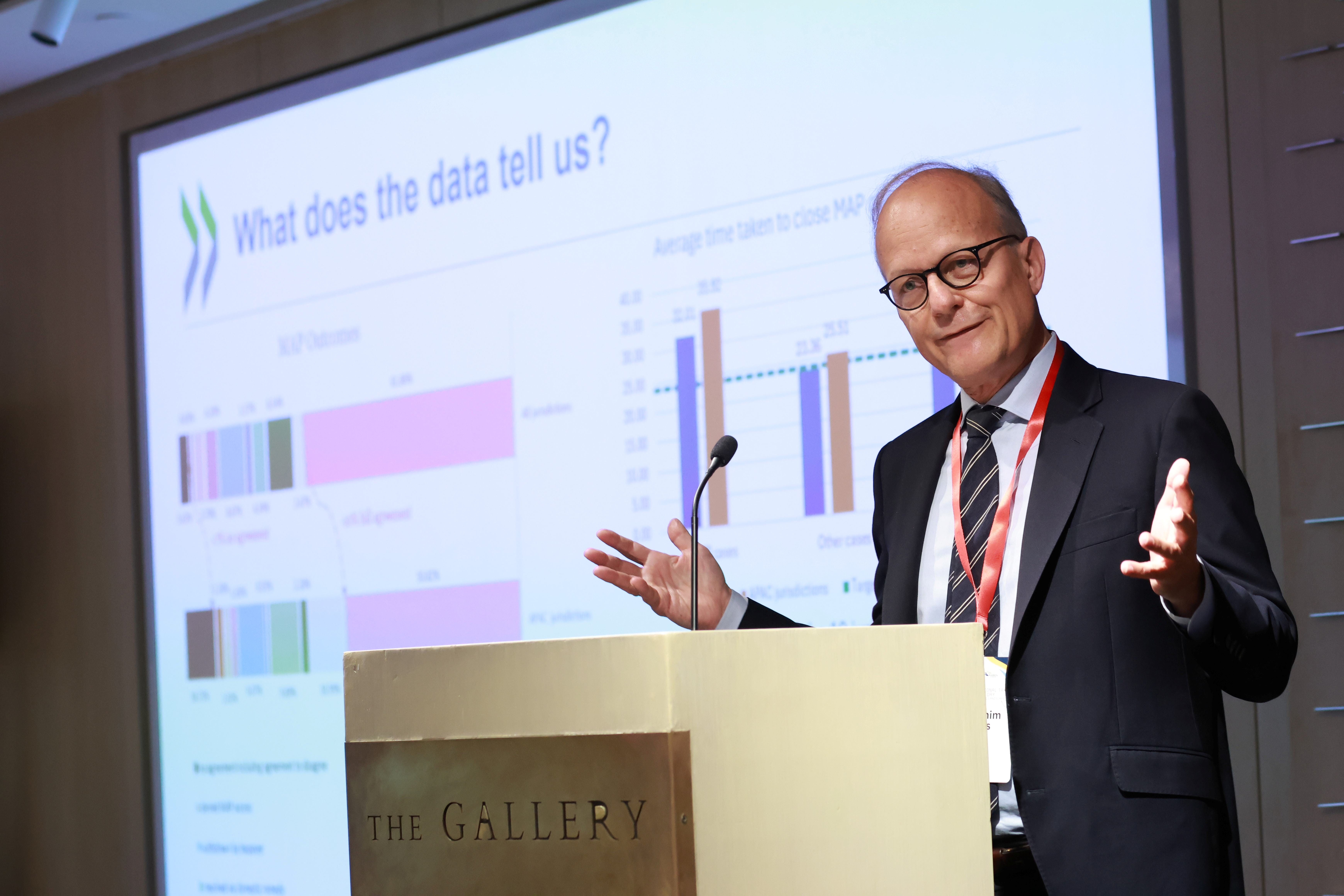 A man in a suit speaks at The Gallery podium before an OECD slide titled "What does the data tell us?"