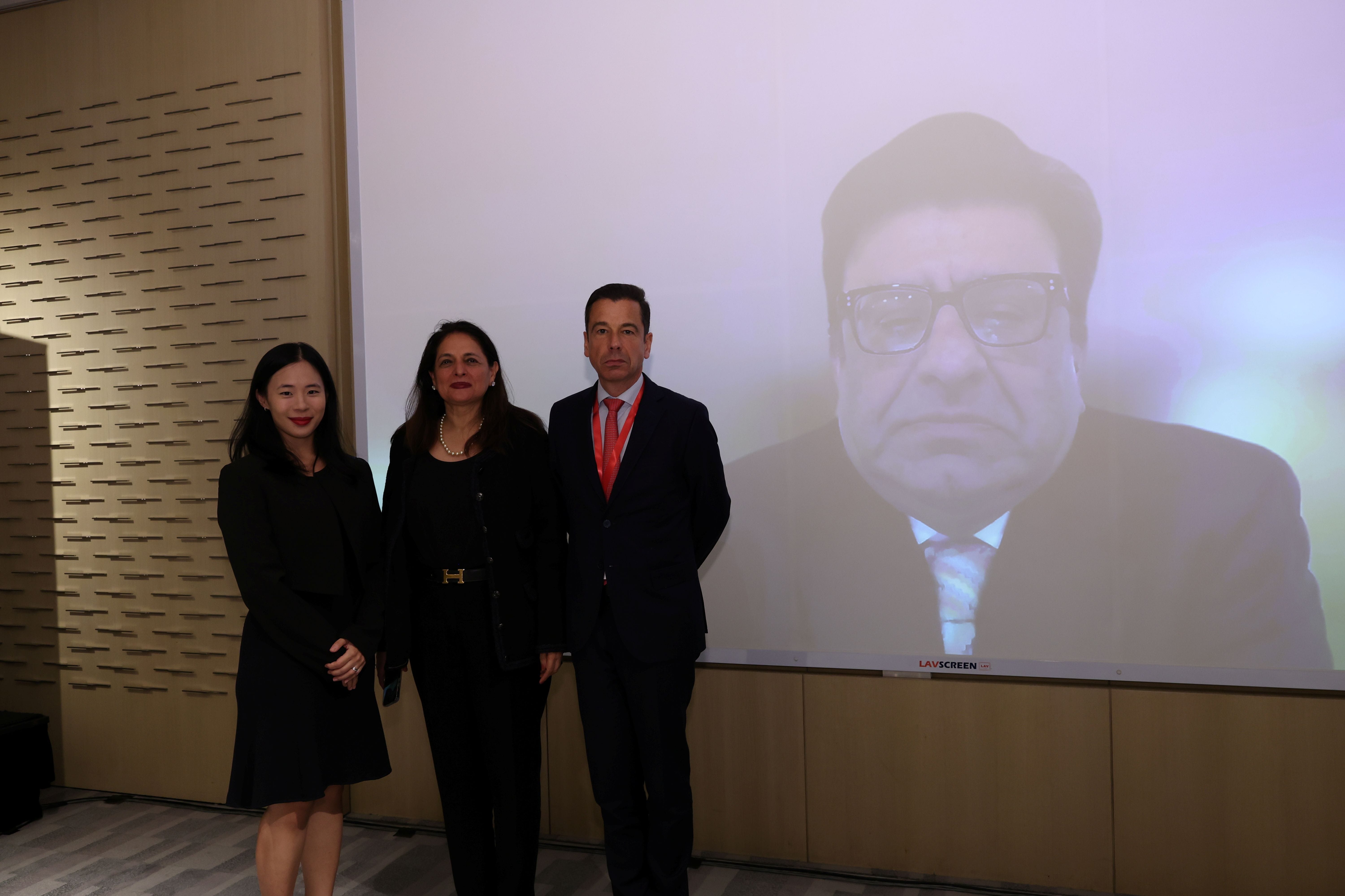 Three people in formal attire stand before a large projection screen showing a man with glasses in a suit.