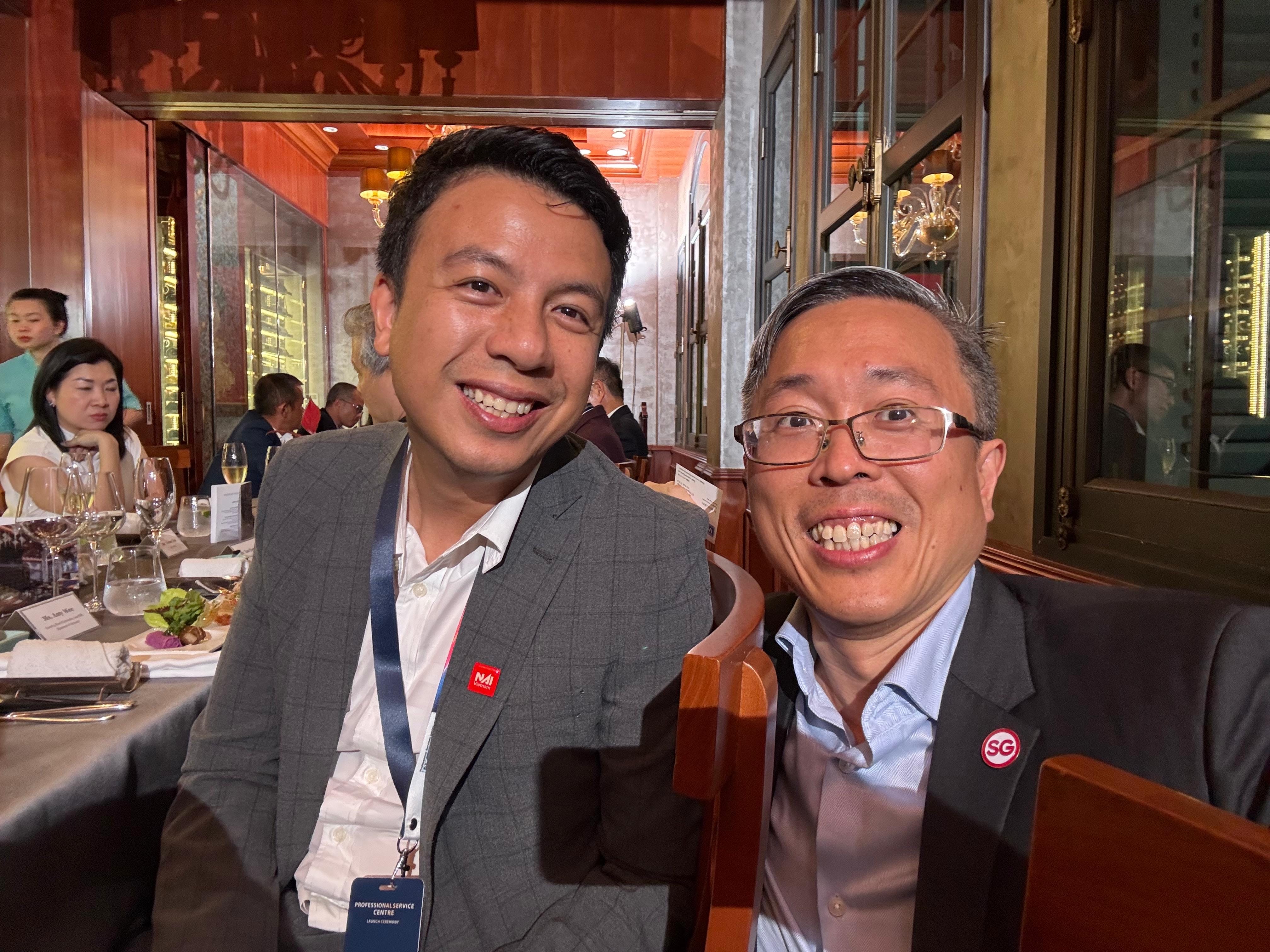 Two smiling men in suits sit together at a formal dinner event inside an elegant restaurant.