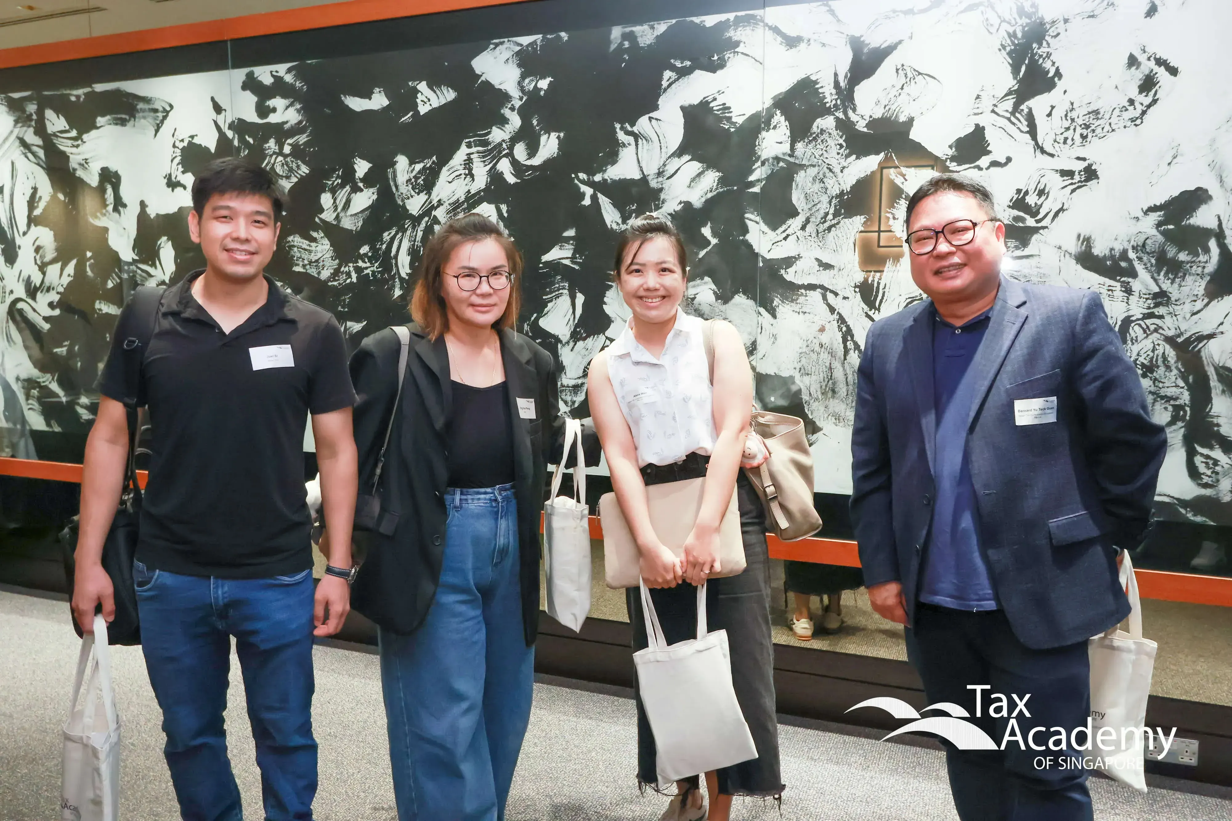 Four people smiling and posing indoors in front of a large black and white abstract mural at Tax Academy Singapore.