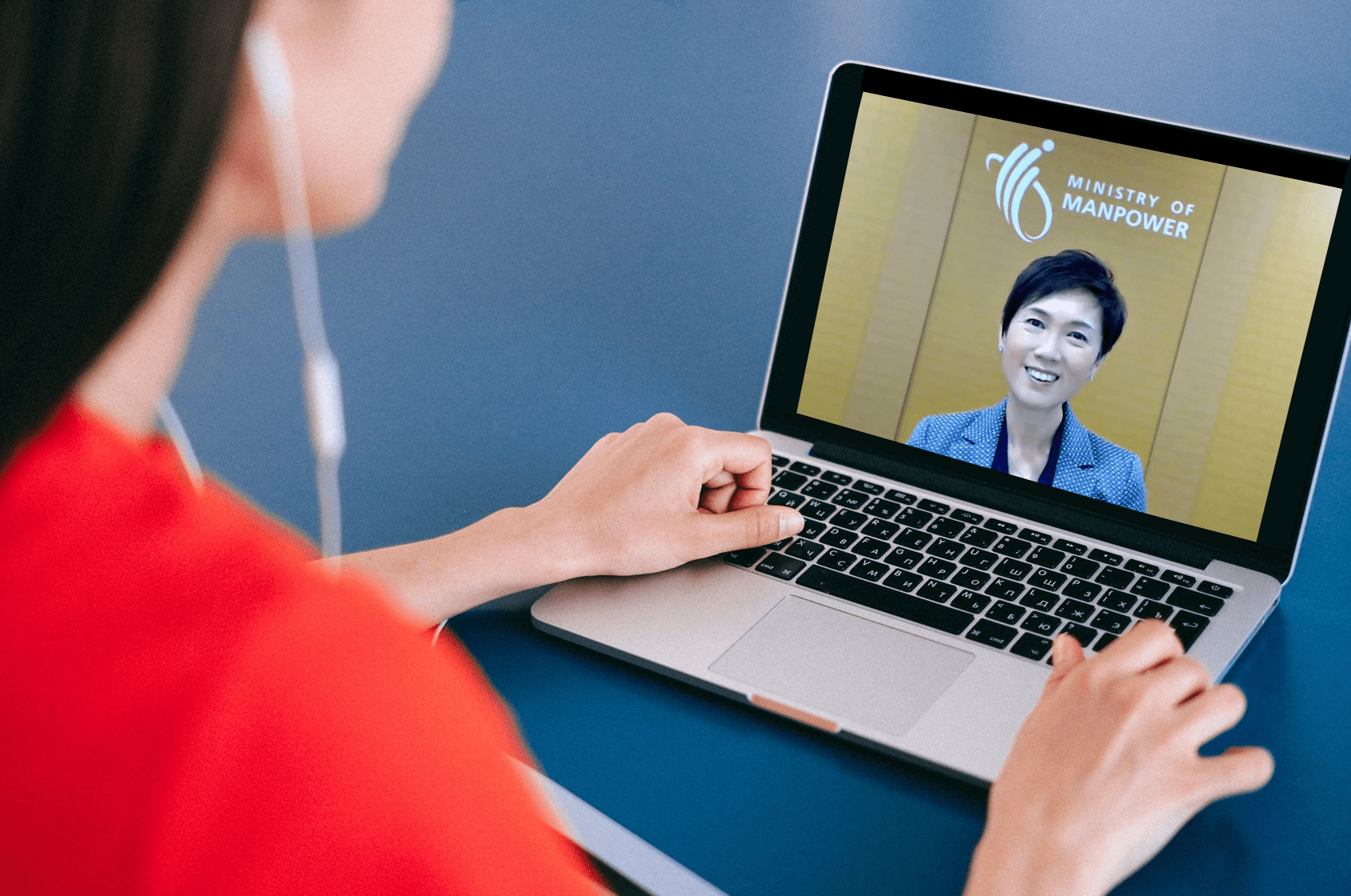 Woman in red top with earbuds watches a Ministry of Manpower video call on a laptop showing a smiling woman.