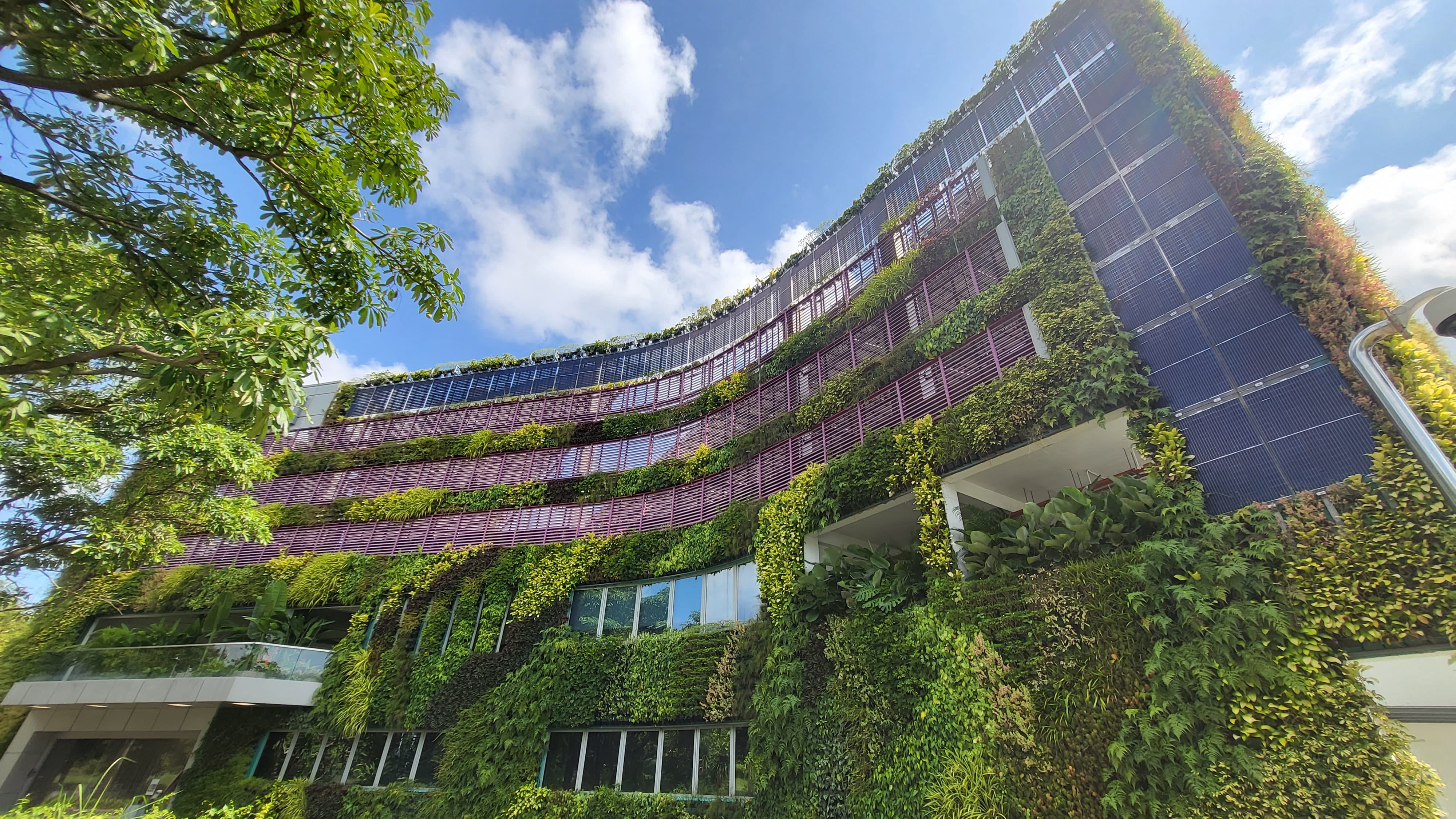 A curved multi-story building covered in lush green vertical gardens and solar panels under a blue sky.