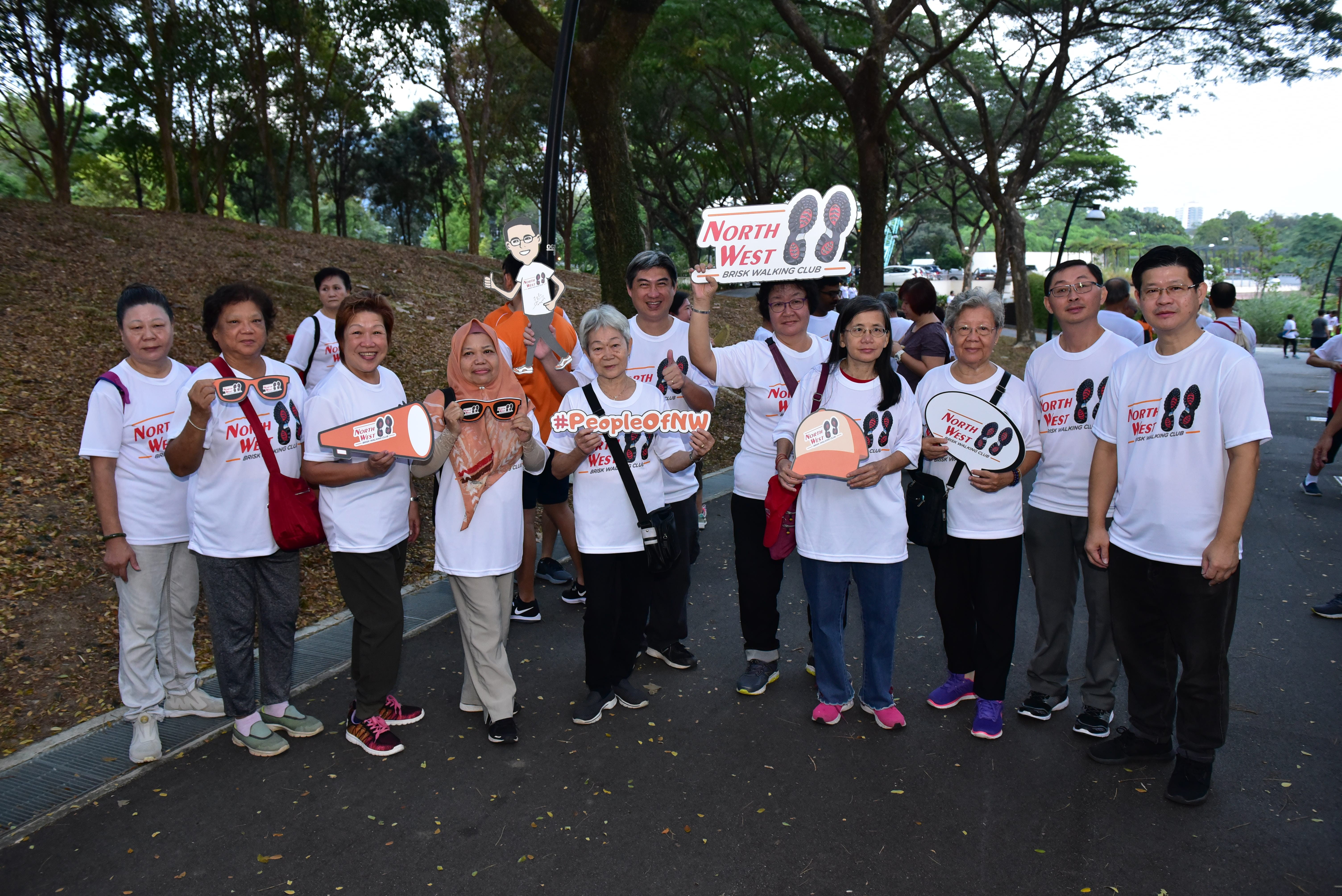 Group of people in white North West Brisk Walking Club shirts posing outdoors holding props and a club sign.