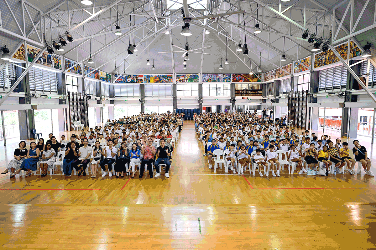 Large group of students and adults seated in rows on a school gymnasium floor with colorful murals on walls.