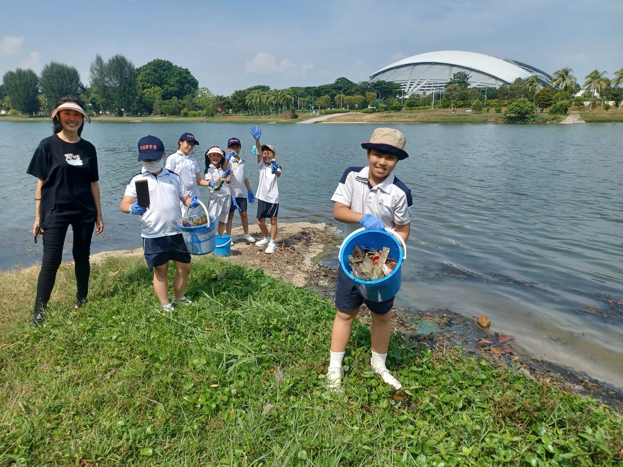 Children in school uniforms hold blue buckets of trash while cleaning up a lakeside area near a large domed stadium.