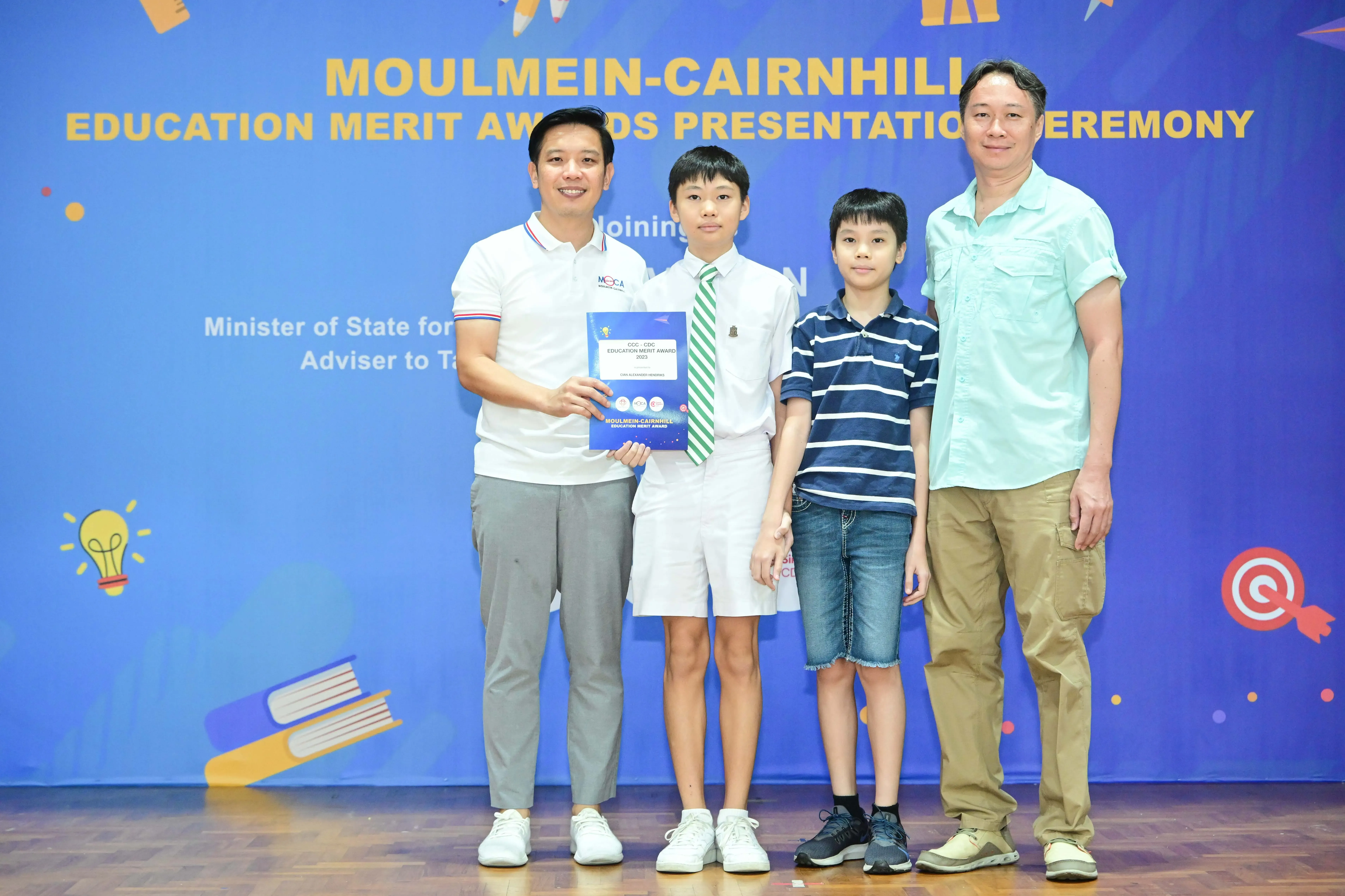 Four people pose at the Moulmein-Cairnhill Education Merit Awards Presentation Ceremony, with a student holding an award certificate.