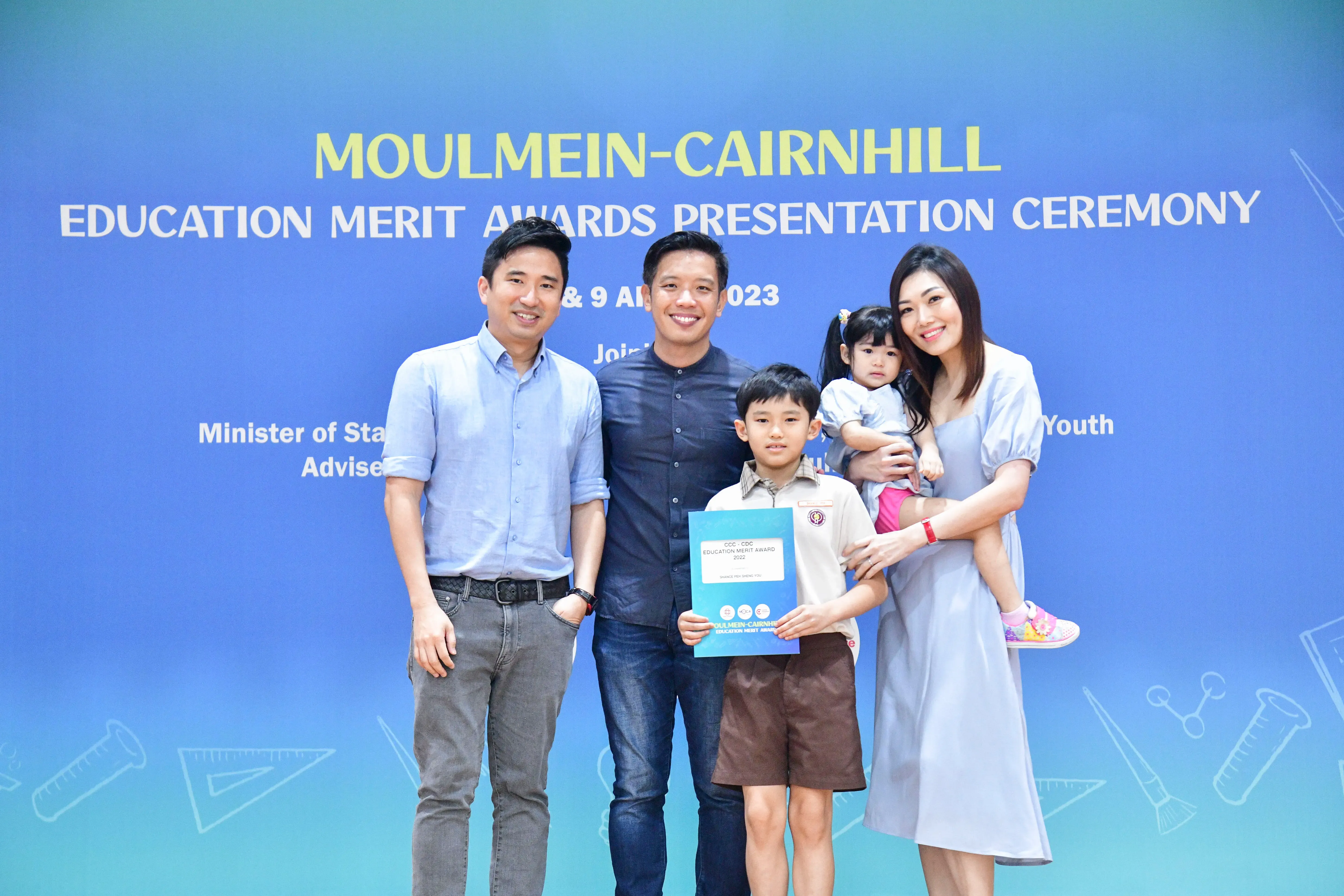Four adults and two children pose at the Moulmein-Cairnhill Education Merit Awards Ceremony 2023, boy holds certificate.